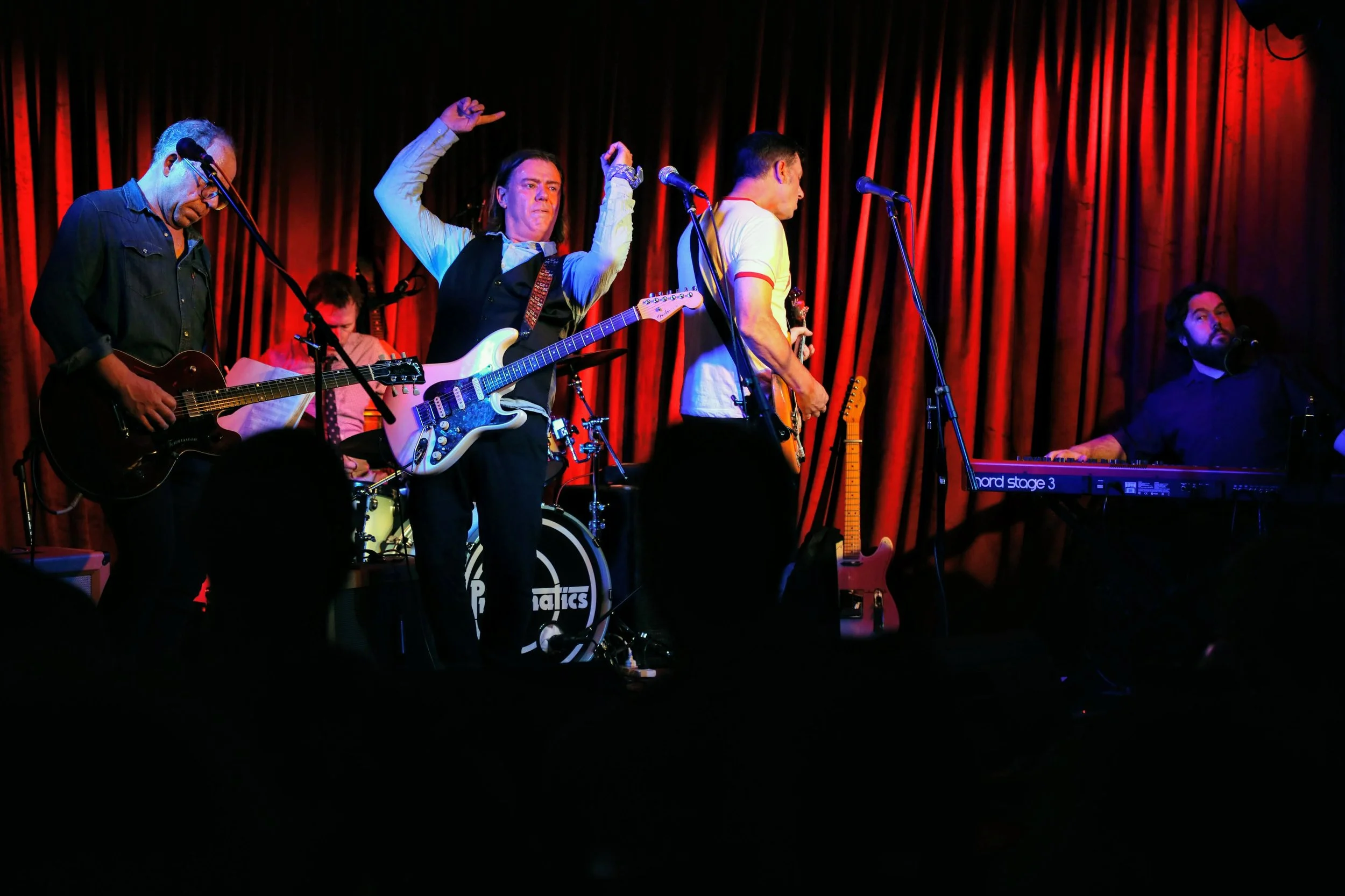 A band performing on stage with red curtains in the background, featuring five musicians playing guitars, drums, keyboard, and singing.