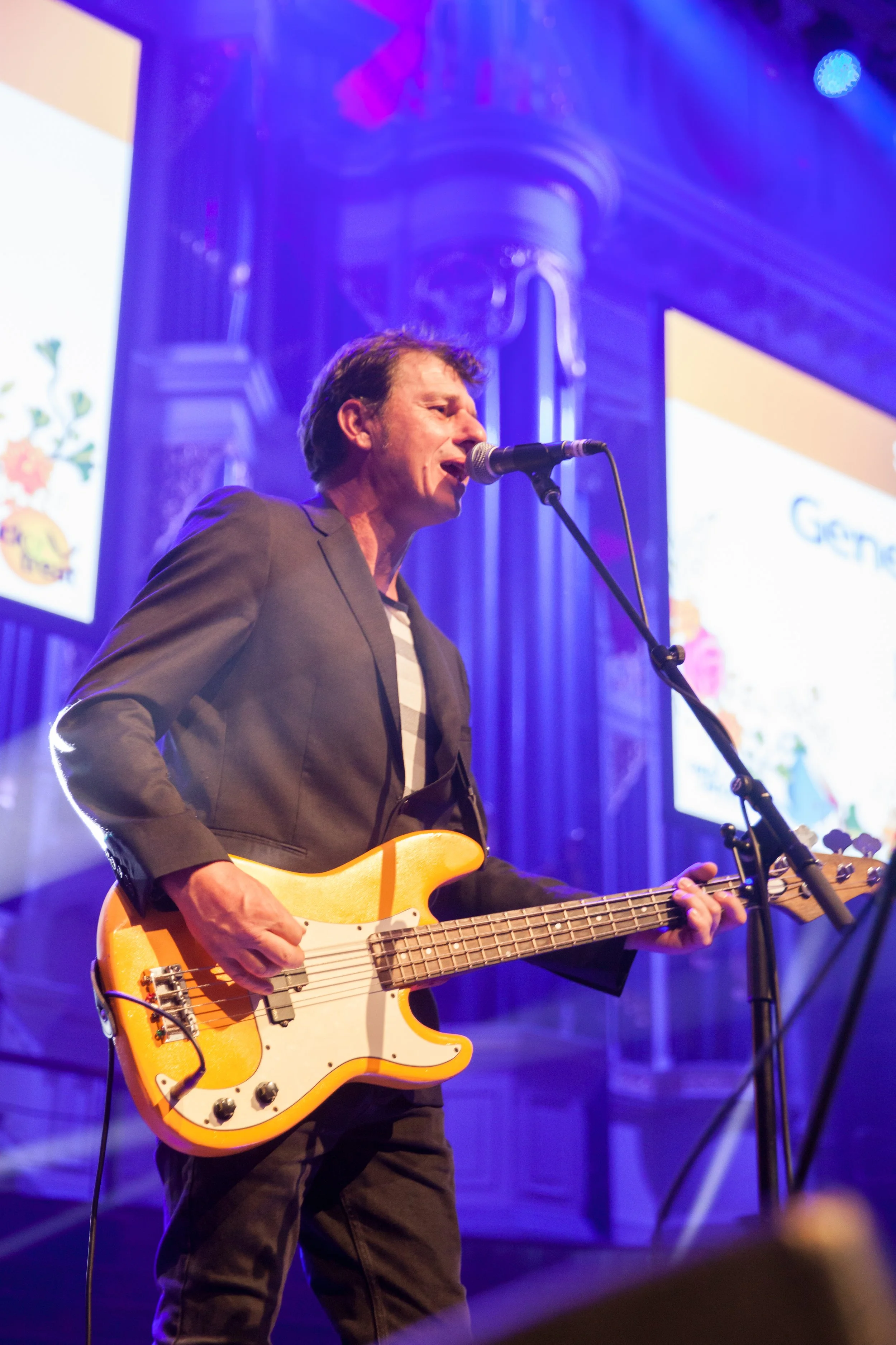 A man performing on stage with an electric bass guitar, singing into a microphone, with large screens and ornate architectural details visible in the background.