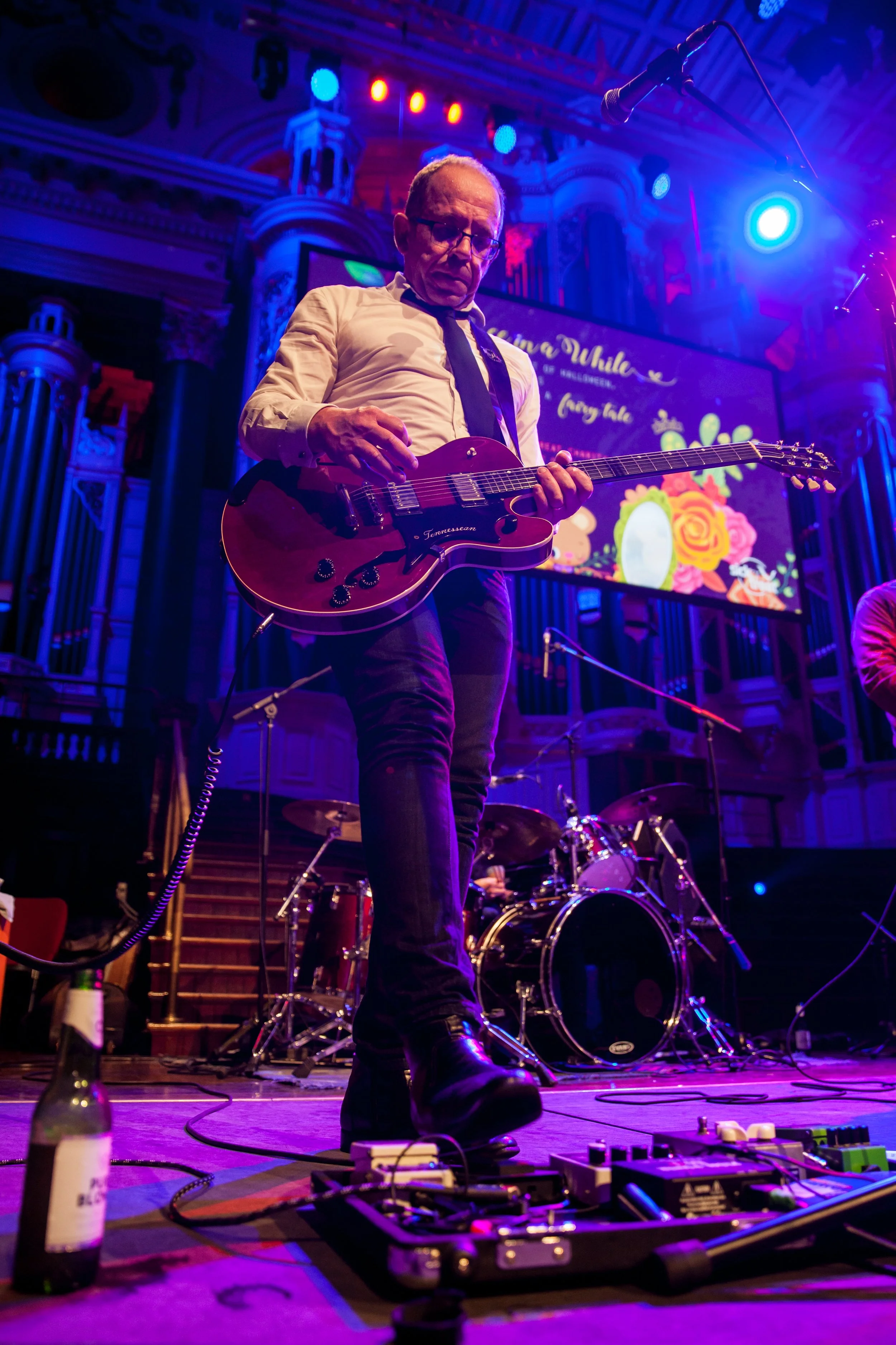 A man playing an electric guitar on stage during a live music performance with colorful stage lighting and a large screen in the background.