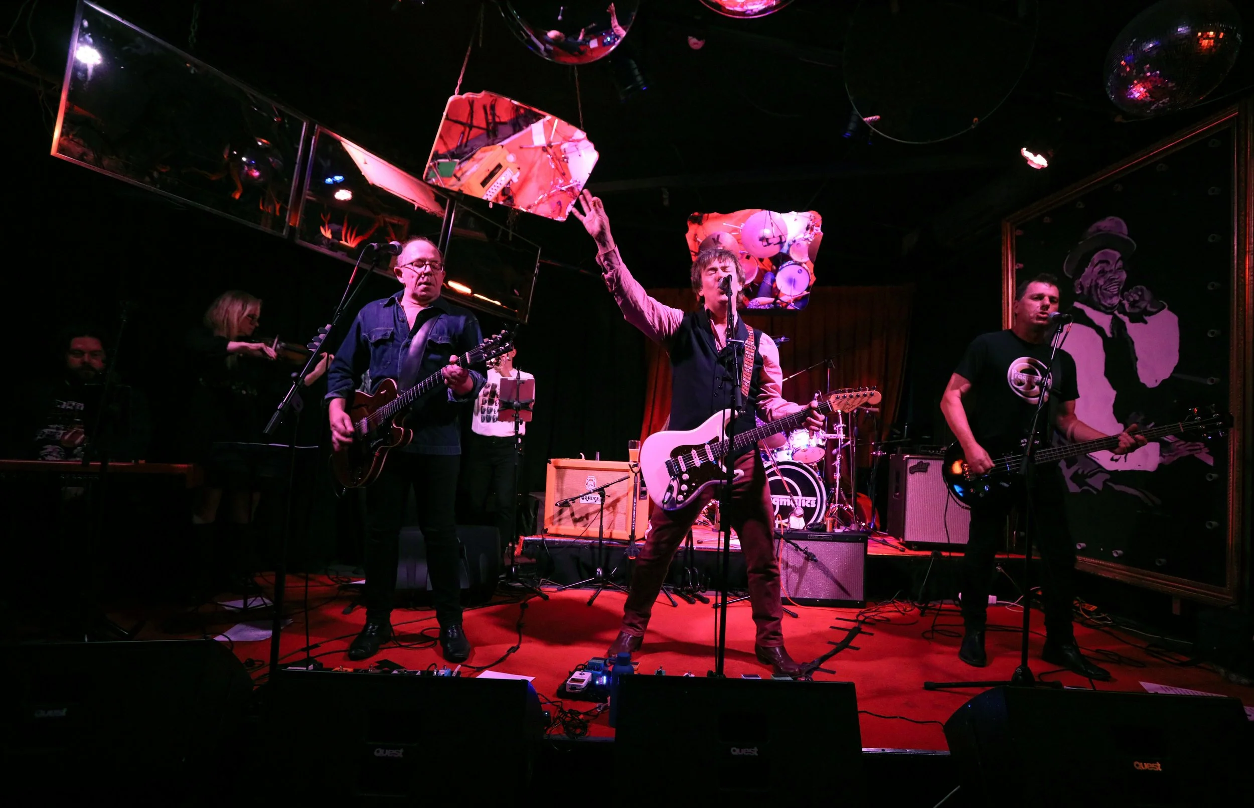 A band performs on stage with five musicians playing guitars and drums, with a woman violist in the background and abstract mirrors and artwork in the background.