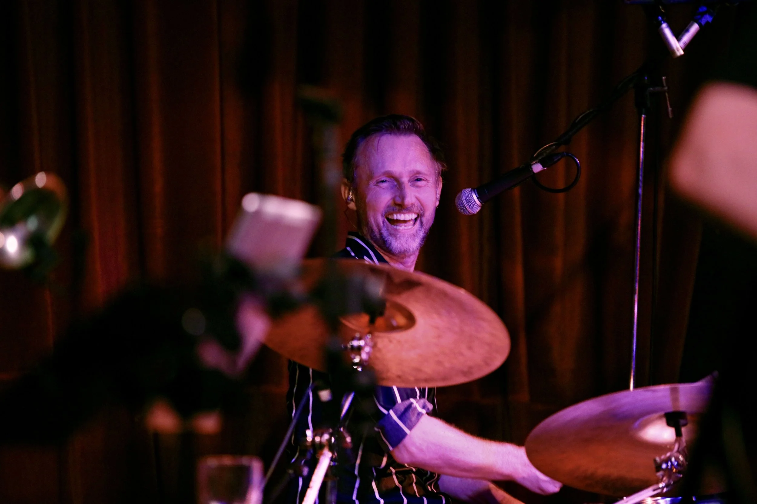 A smiling man playing drums at an indoor music performance, with a microphone nearby, in a dimly lit venue with dark curtains in the background.
