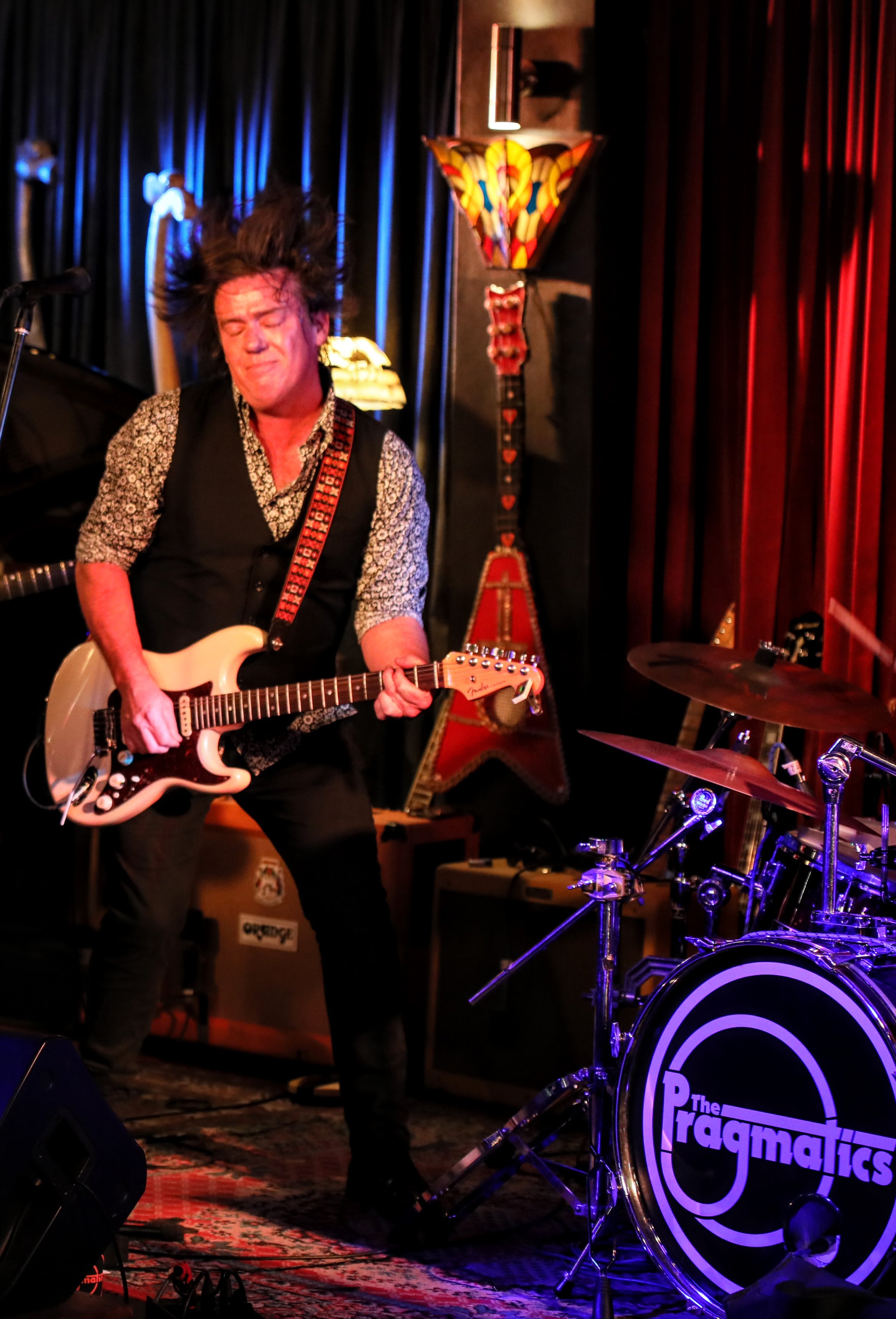 A man playing an electric guitar on stage with a band. The stage has blue and red curtains, and there are guitars and amplifiers in the background. The drum set has a logo that reads 'The Pragmatiques.' The man has dark hair and is wearing a patterne