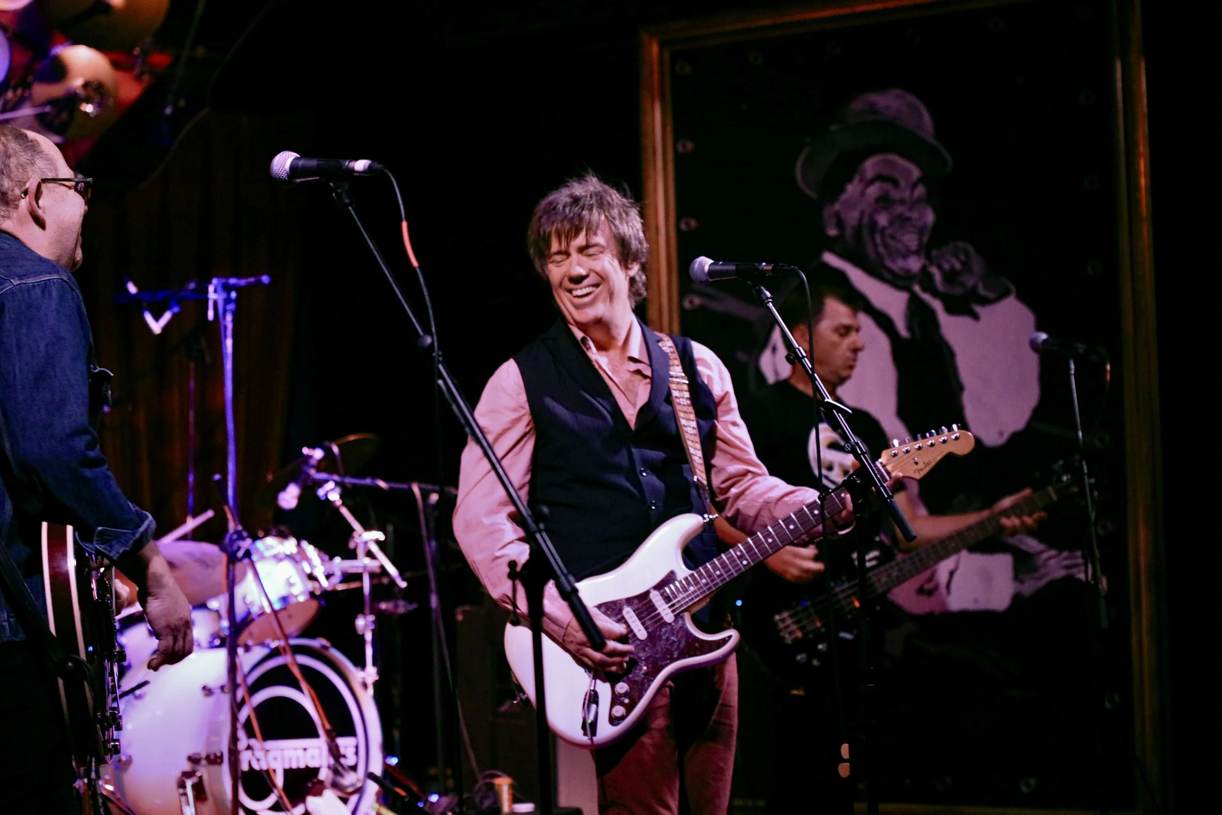Four musicians perform on stage, with the central figure playing an electric guitar and smiling, dressed in a pink shirt and black vest, in front of a black and white portrait of an elderly man with a hat.