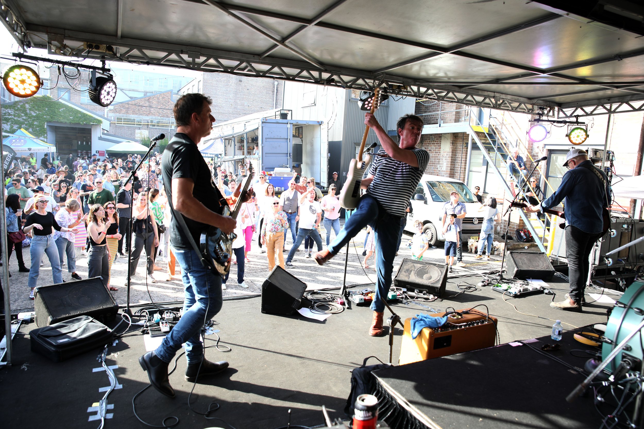 A band performing on stage at an outdoor concert with a crowd dancing in front and people watching in the background.