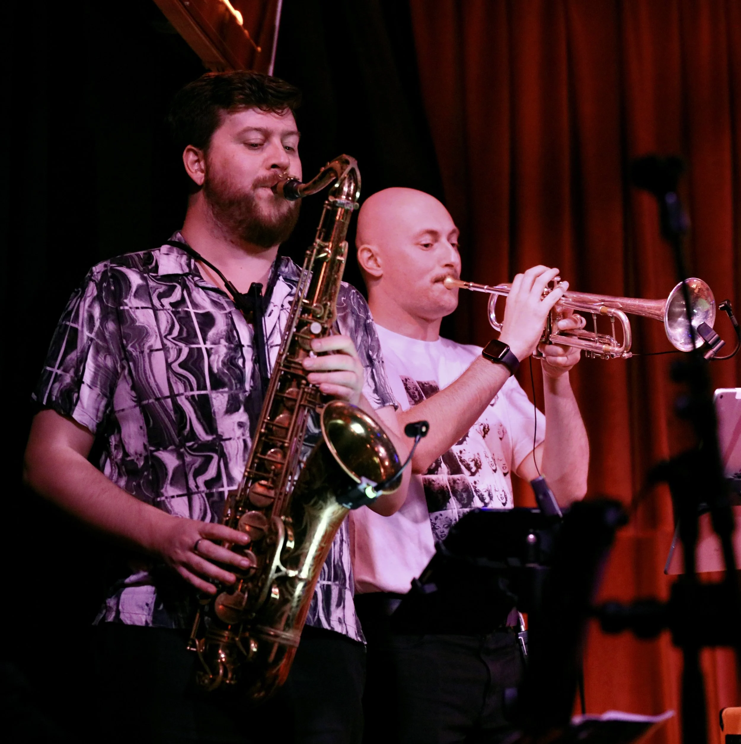 Two jazz musicians playing saxophone and trumpet on stage with red curtains in the background.