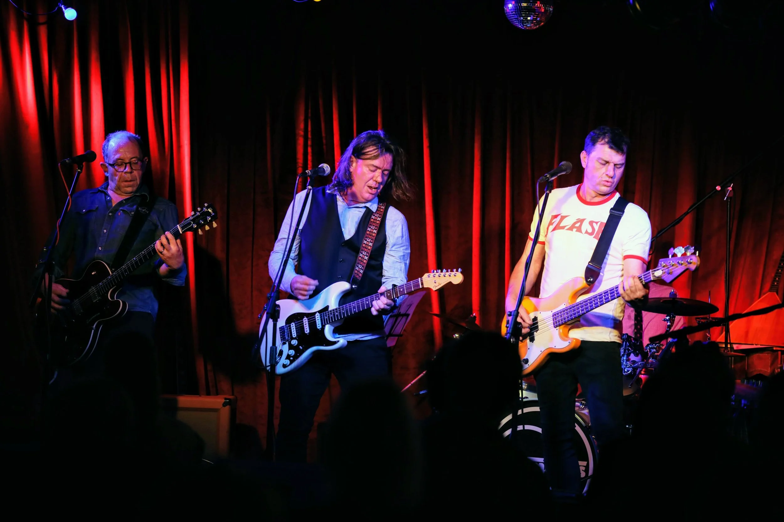 Three musicians performing on stage with a red curtain background. The musician on the left is playing an electric guitar, the middle musician is singing and playing an electric guitar, and the right musician is playing a bass guitar. All are focused