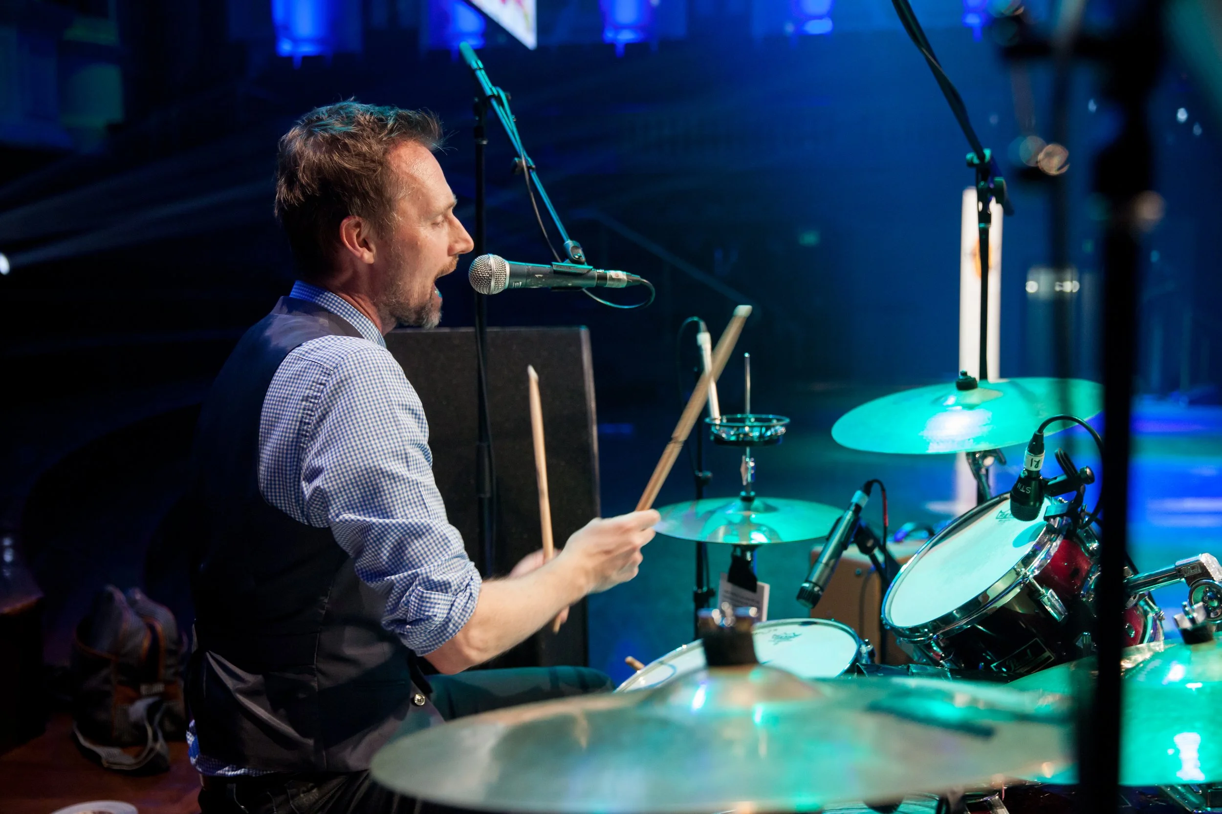 A man playing the drums, singing into a microphone on a stage with blue lighting.