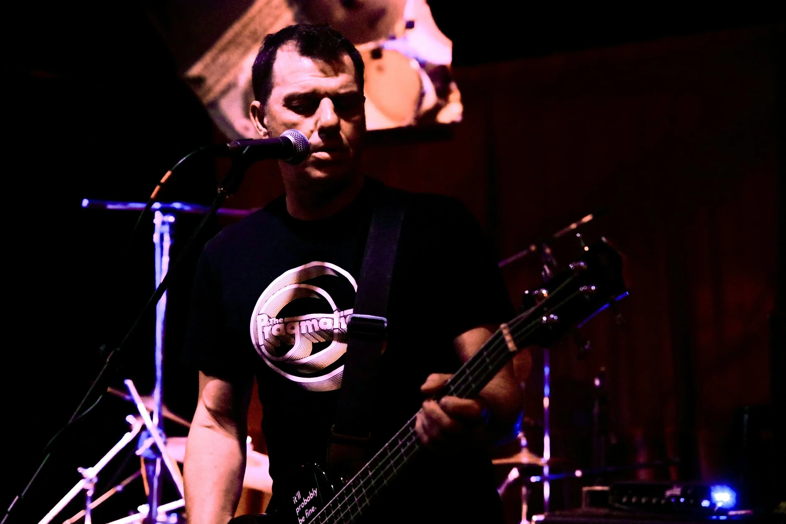 A male musician playing an electric guitar and singing into a microphone on a stage with drum set and sound equipment in the background, in a dimly lit venue.