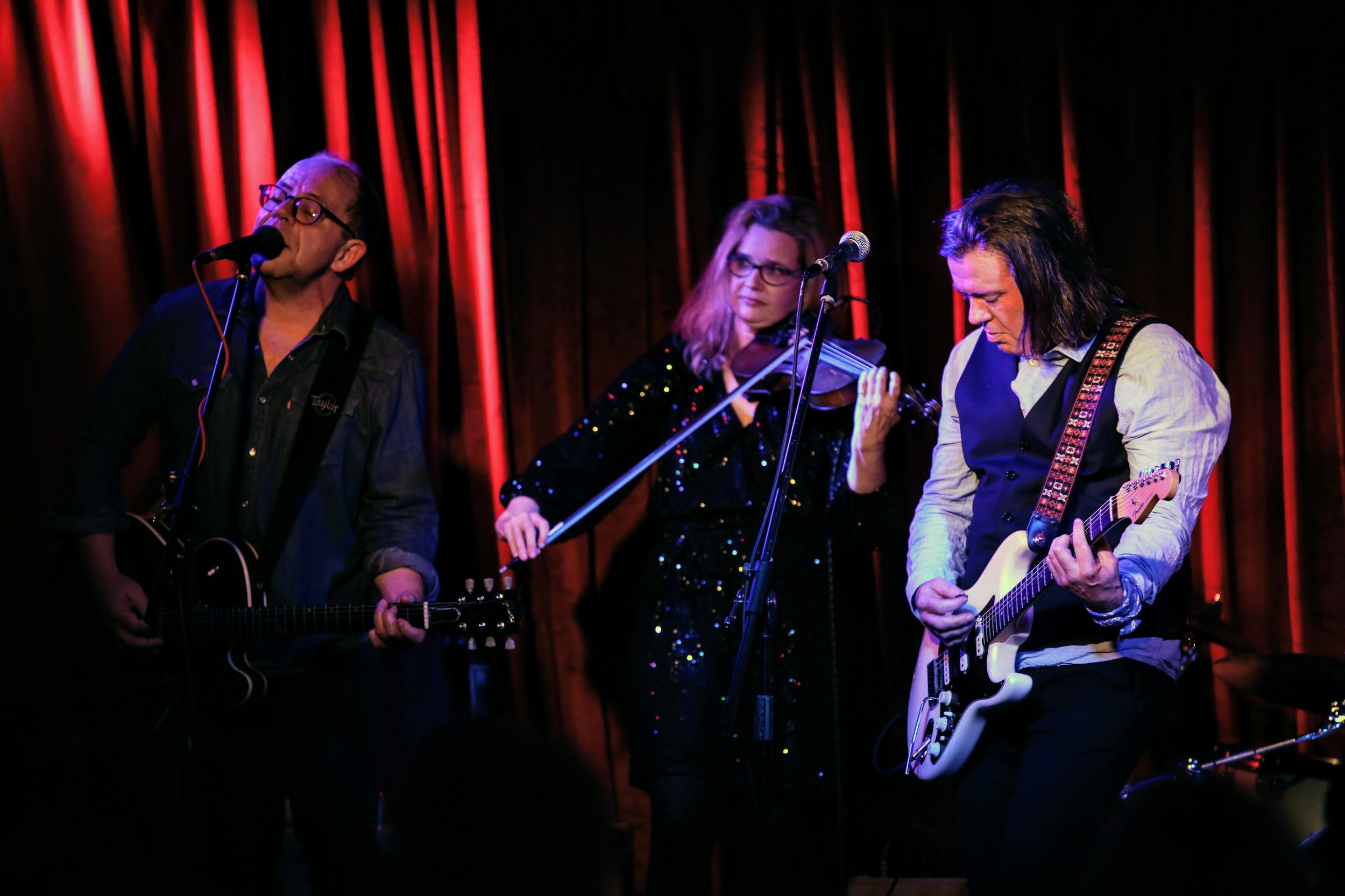 Three musicians performing on stage: a man singing and playing guitar, a woman playing violin, and another man playing electric guitar, with red curtains in the background.