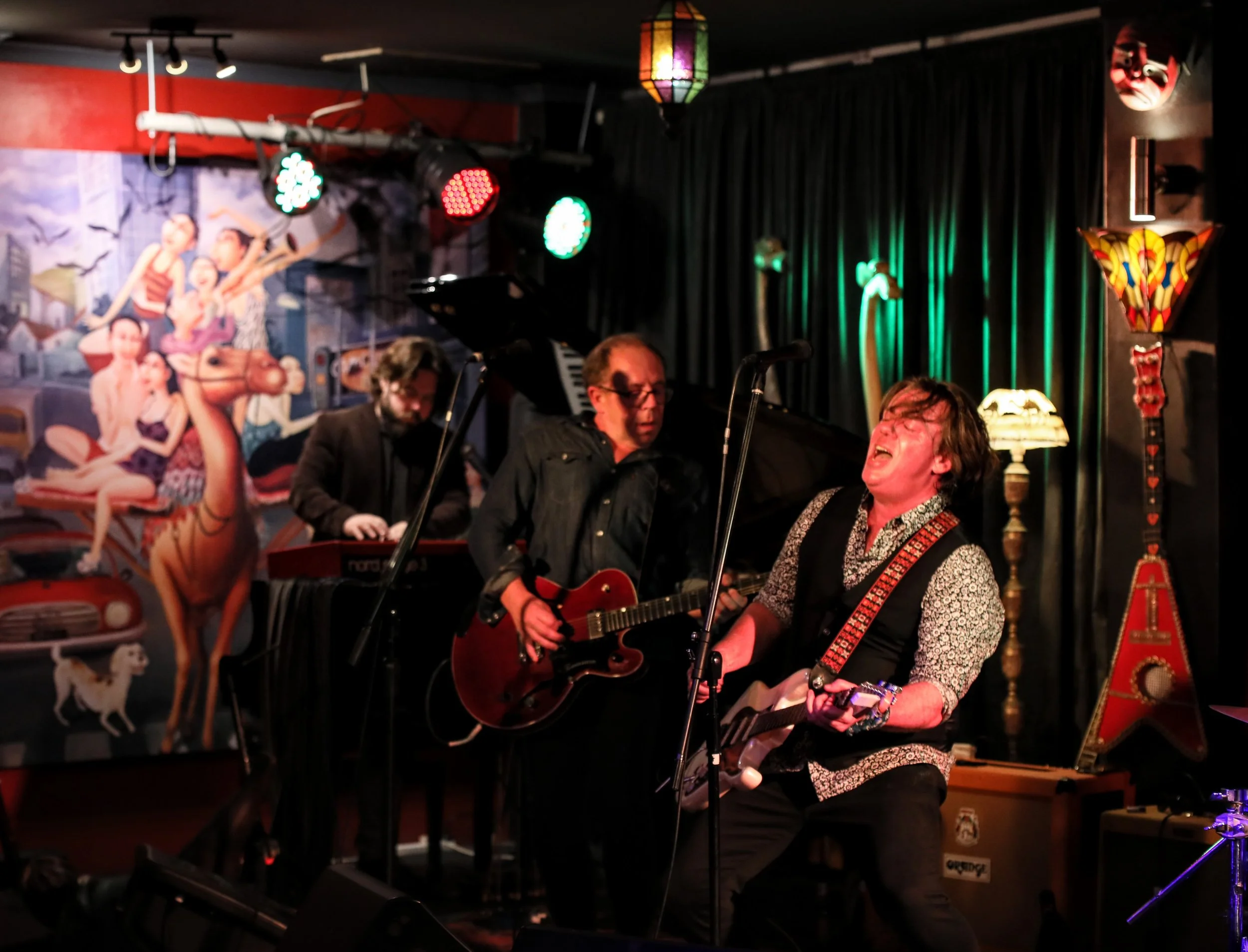 Three musicians performing on stage at a bar or club, one playing guitar and singing passionately, another playing an electric guitar, and a third on keyboard, with a colorful mural and decorative lamps in the background.