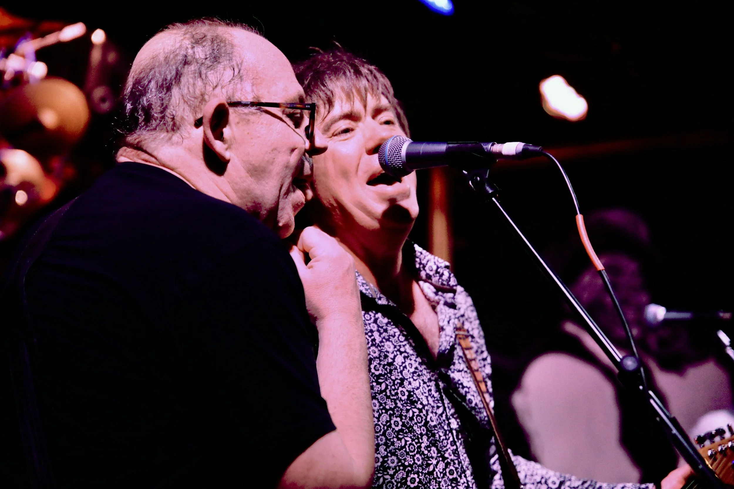 Two men singing into a microphone on stage at a concert, with one playing a guitar in the background.