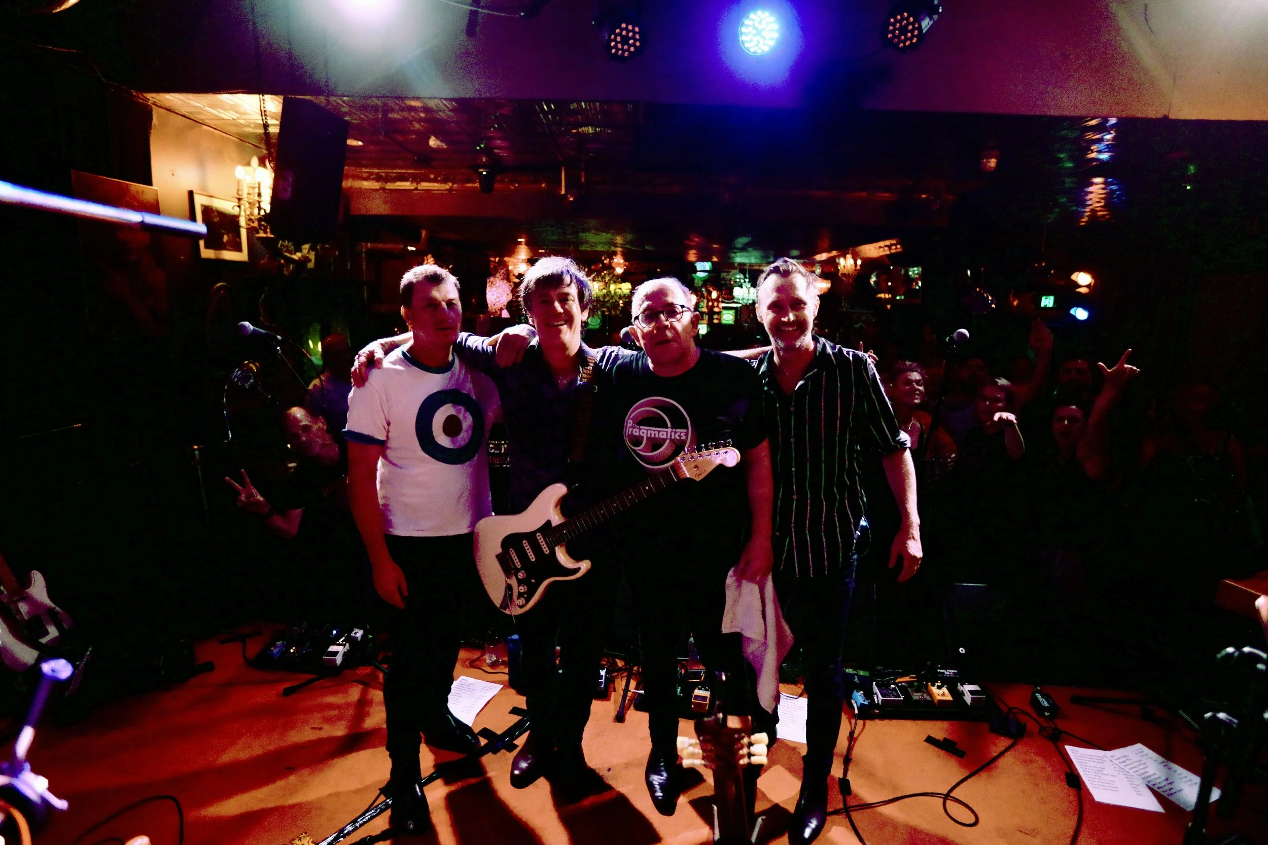 A group of four men on stage with musical instruments, celebrating after a performance, in a venue with an audience in the background.