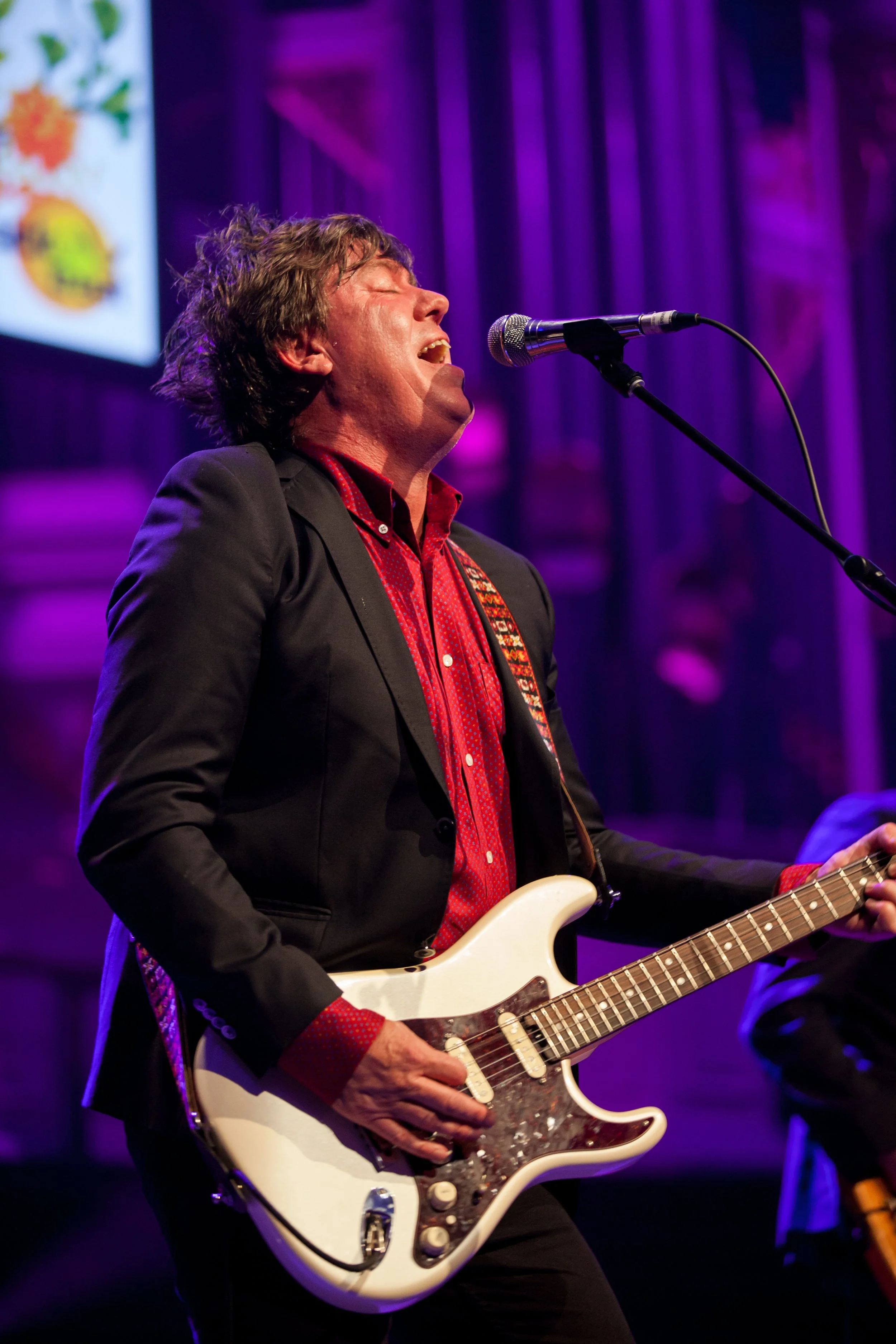 A man passionately singing and playing an electric guitar on stage with purple lighting.