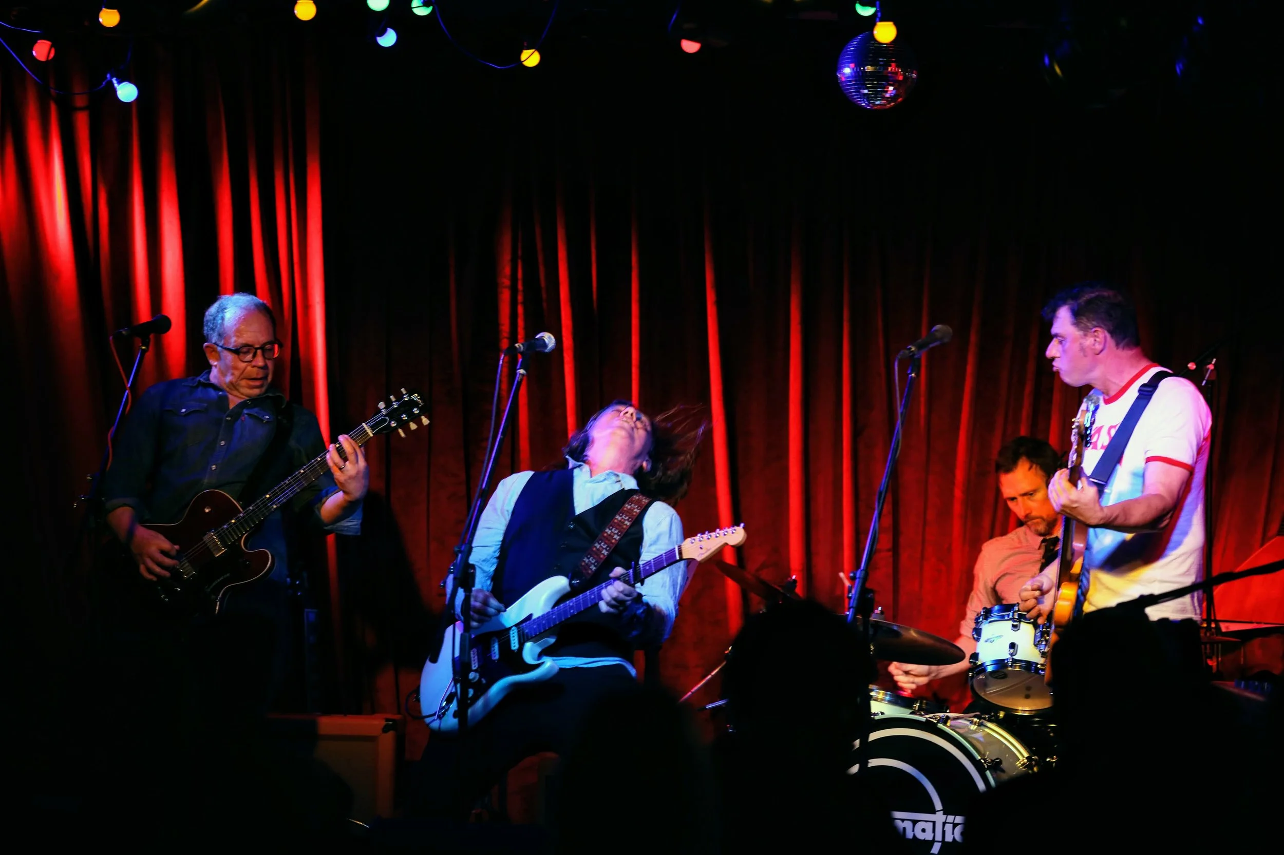 A band performing live on stage with a red curtain backdrop, including a guitarist, a vocalist, a drummer, and another guitarist, with stage lights and a disco ball overhead.