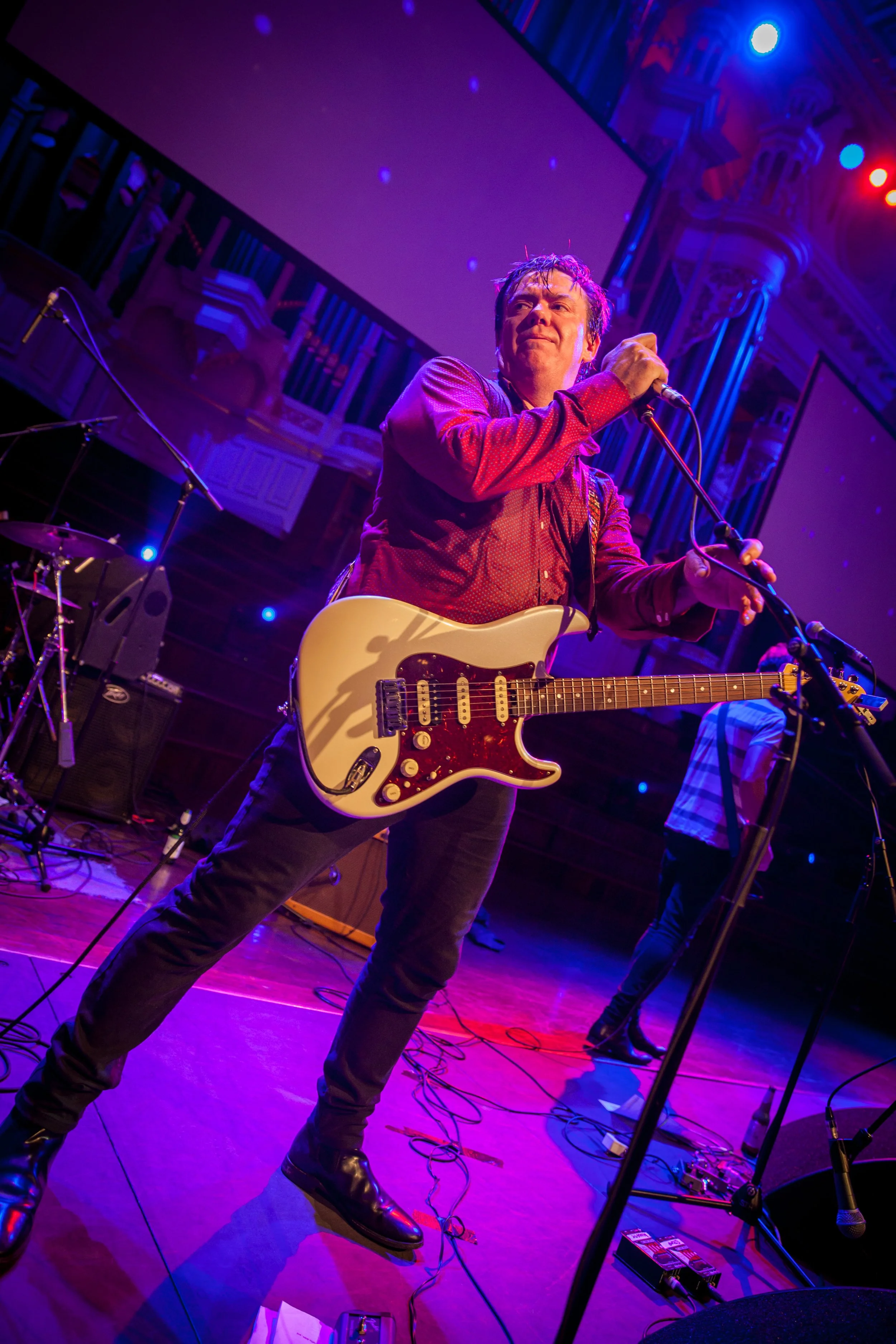A male musician on stage holding a white electric guitar and a microphone during a live performance, with colorful stage lighting and a backdrop with a large screen.