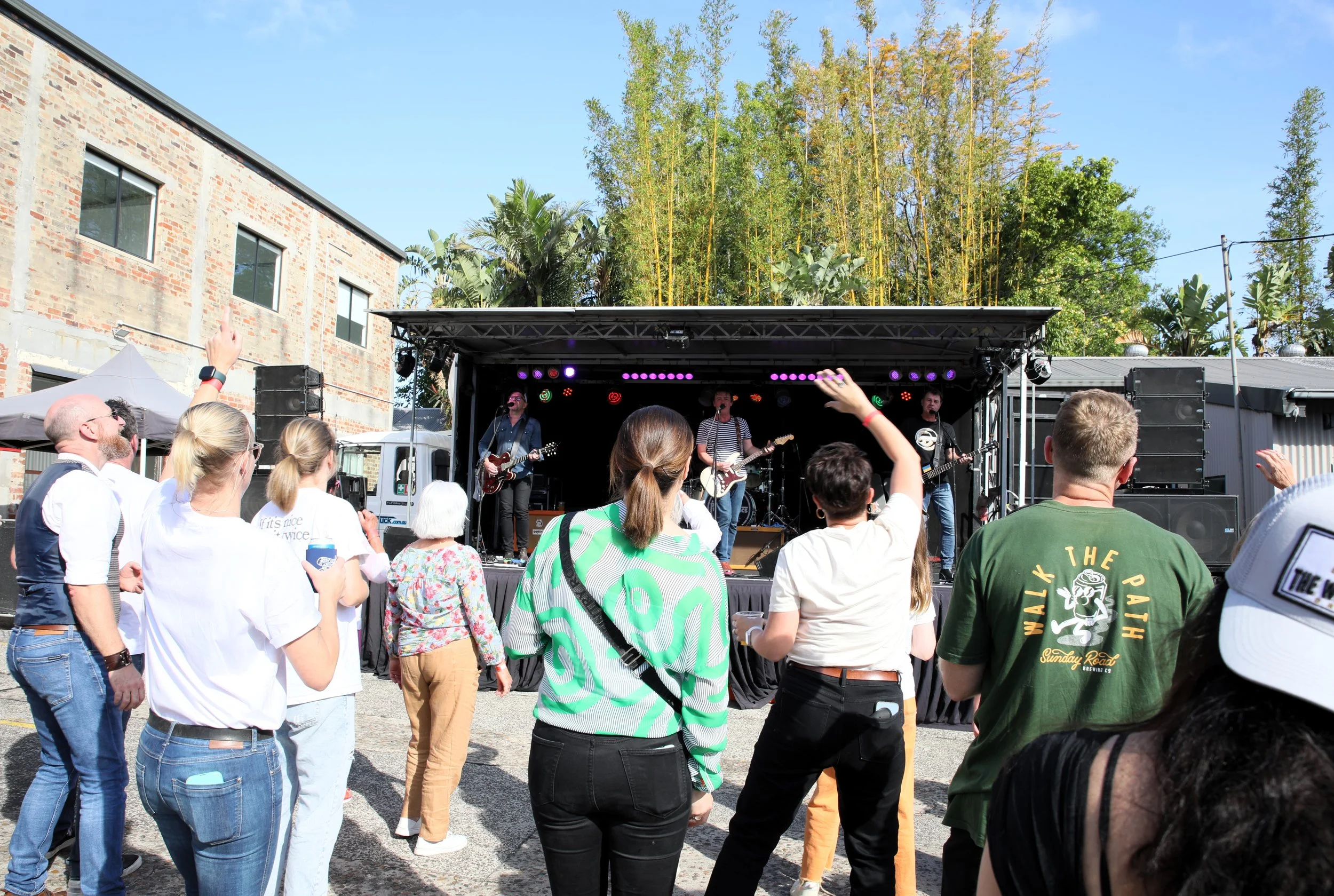 People enjoying a live outdoor concert with a band playing on a stage, surrounded by trees and buildings.