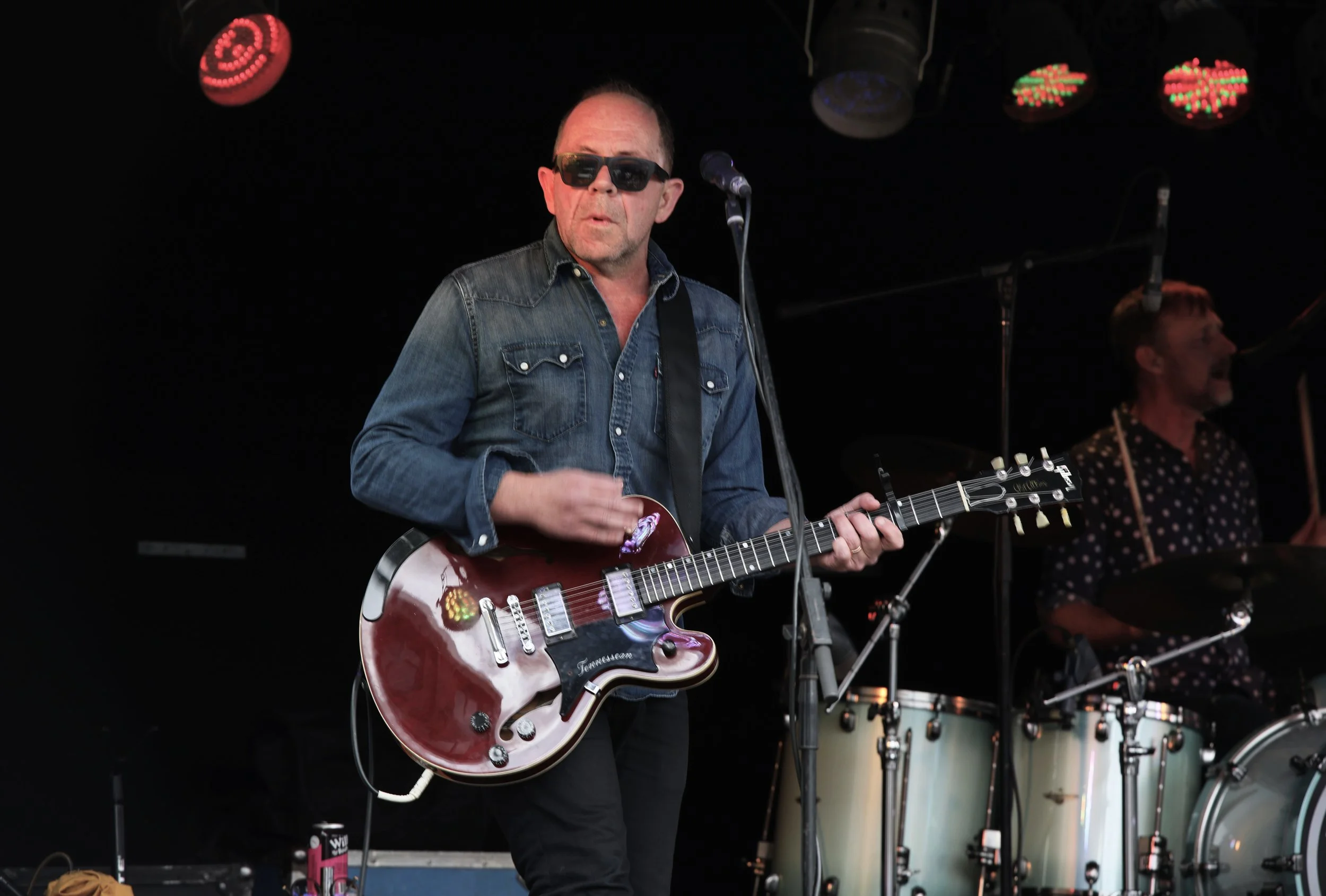 A man wearing sunglasses and a denim shirt playing an electric guitar on stage during a live music performance, with a drummer in the background.