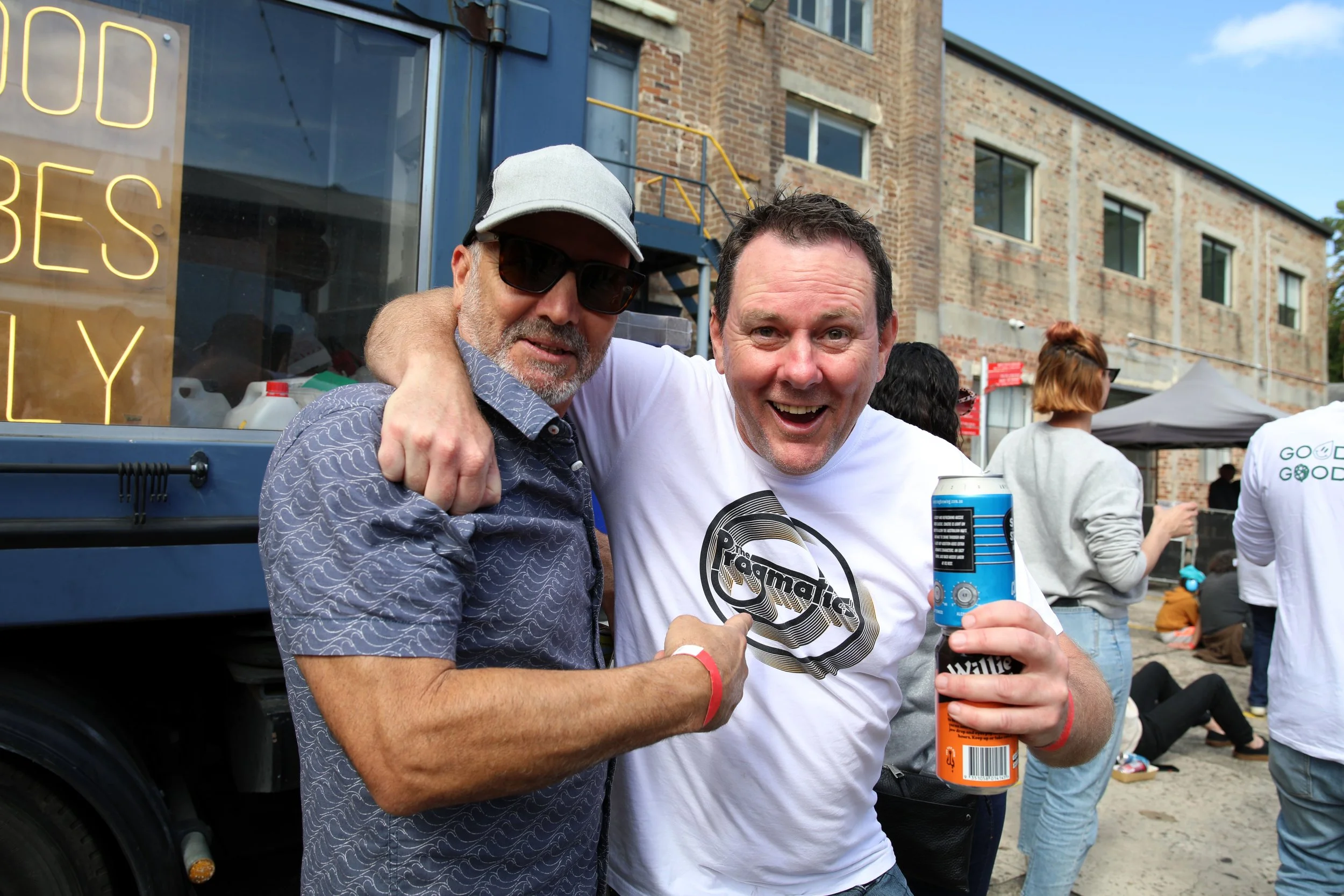 Two men smiling and posing together at an outdoor gathering or event, with a food truck or vendor stand in the background, one man wearing sunglasses and a cap, the other holding a beverage can.