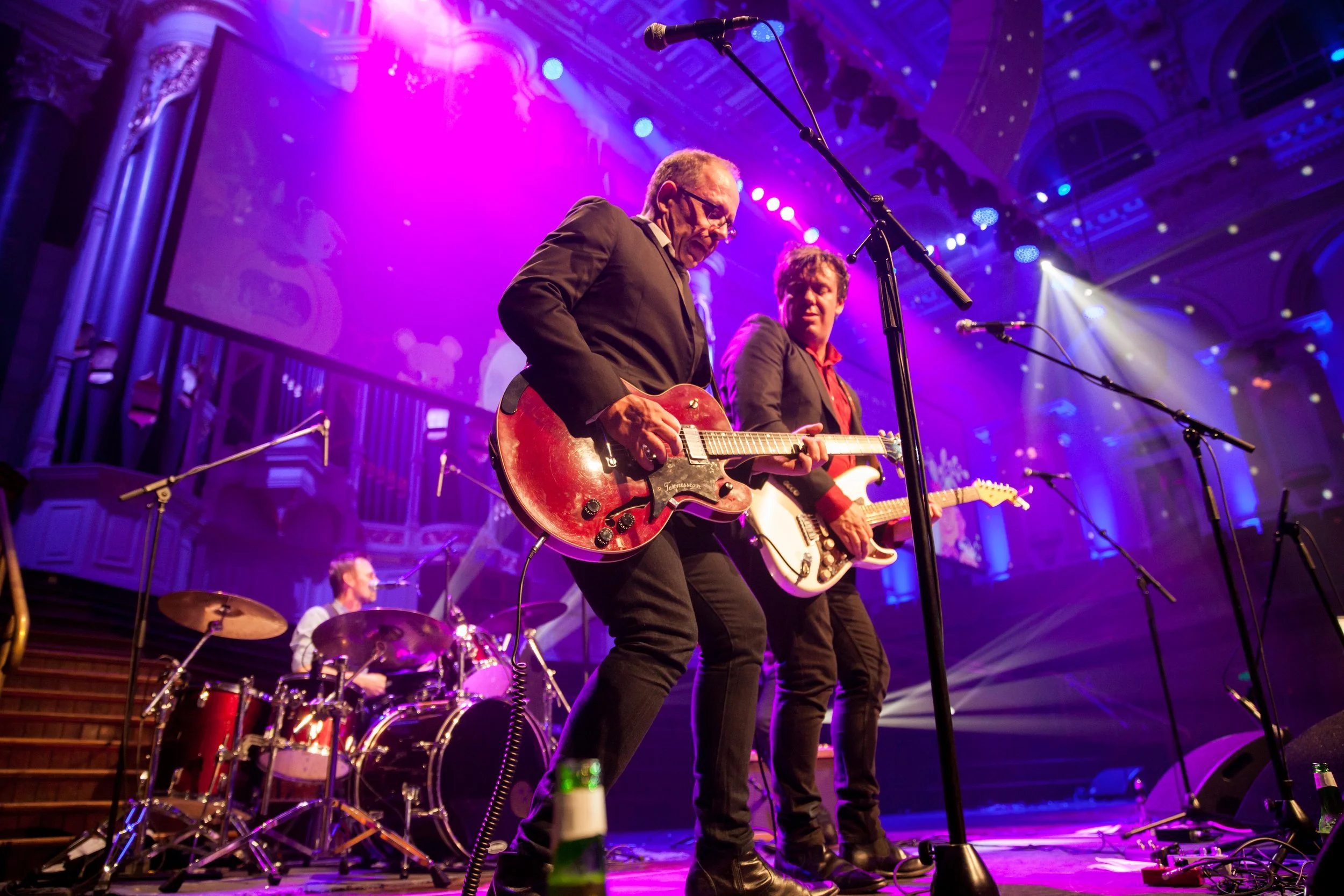 A band performing on stage with purple and blue lighting, featuring a drummer, two guitarists, and vocal microphones.