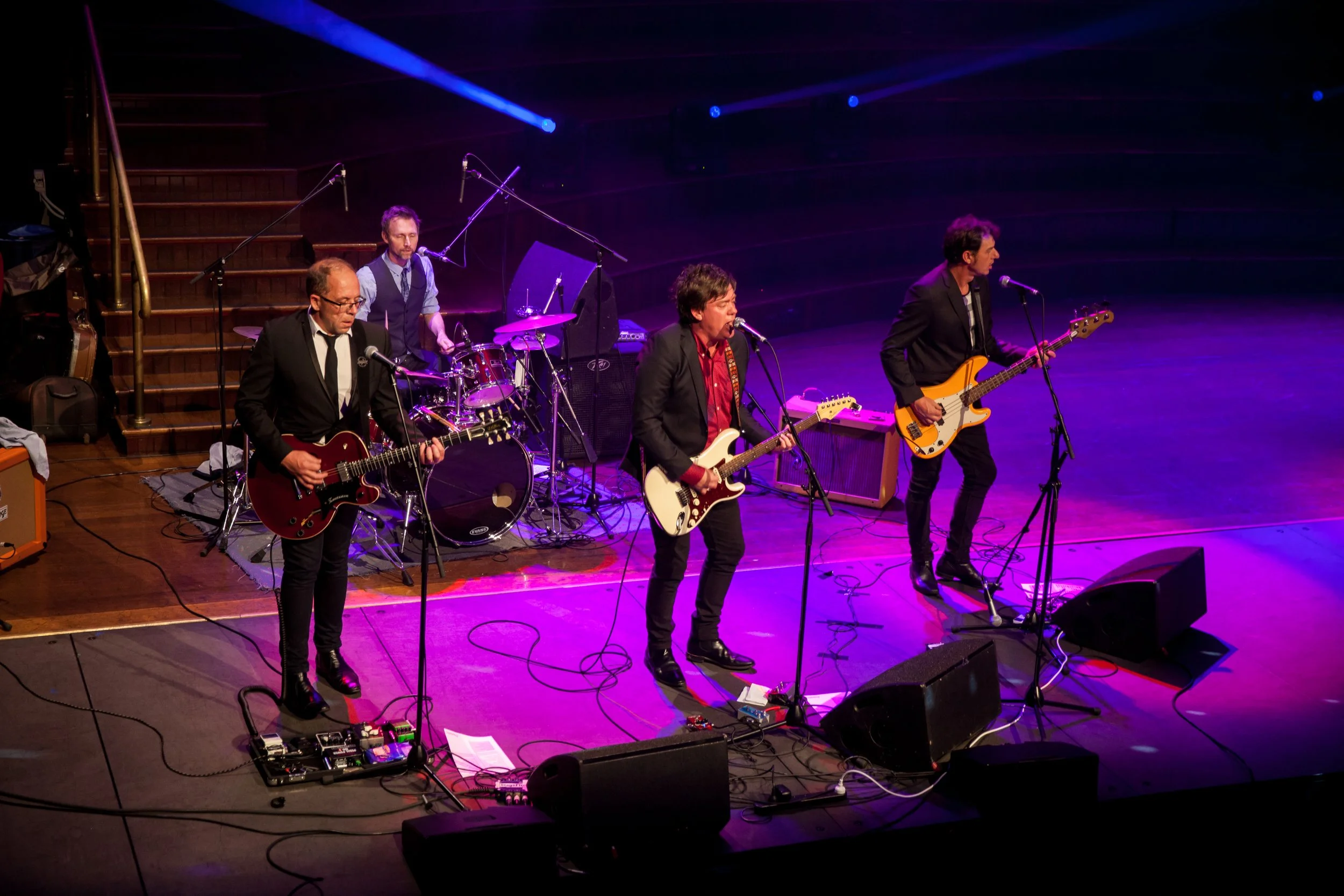 A band performing live on stage, with four male musicians playing guitars and drums under purple and blue stage lighting.