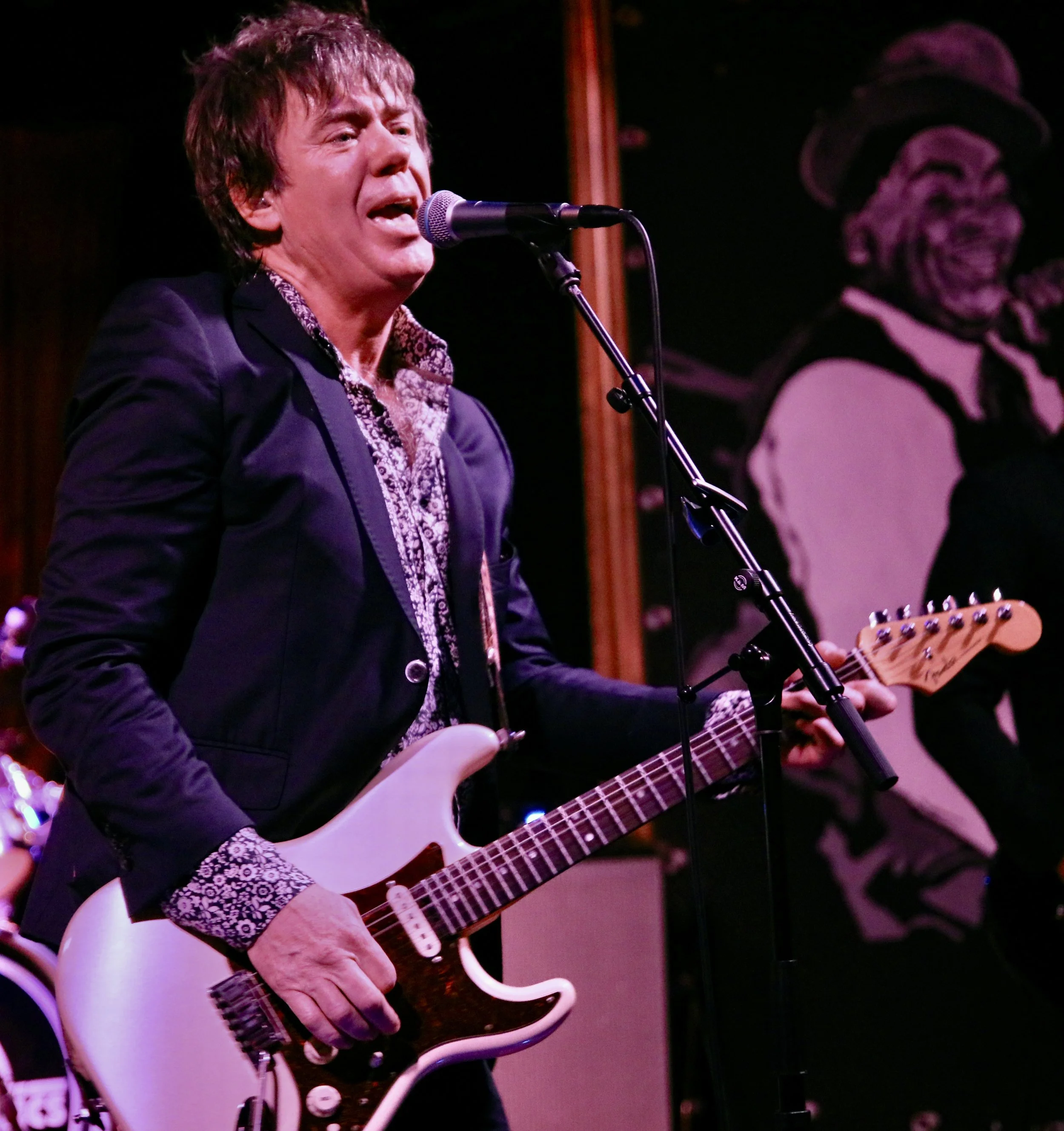 A man playing an electric guitar and singing into a microphone on stage with a portrait of a singer in the background.
