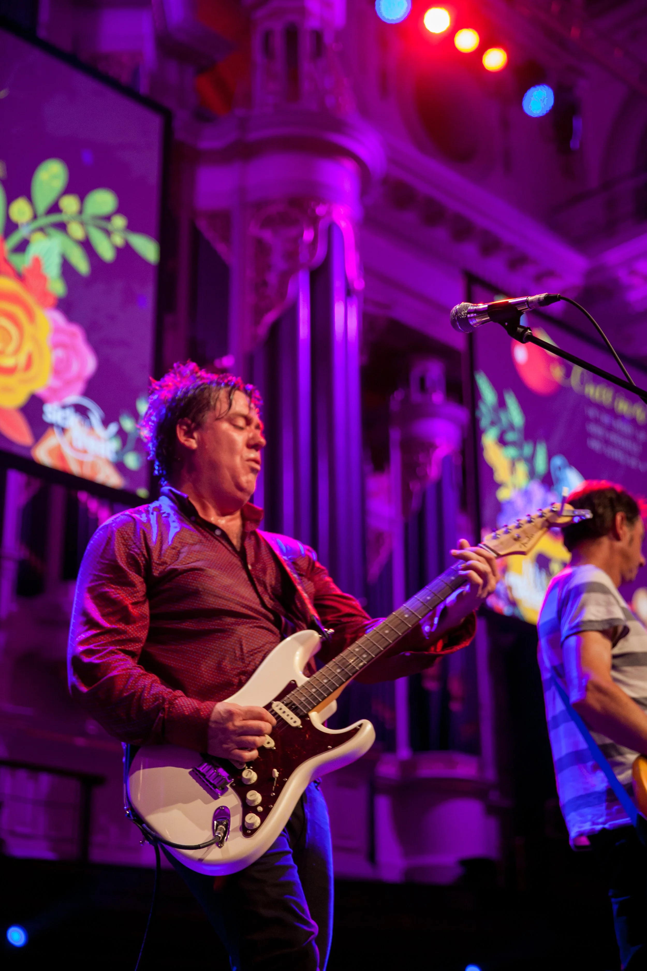 Musicians performing on stage with colorful lighting, one playing an electric guitar and another playing a guitar, with a decorative backdrop and a large screen displaying graphics.