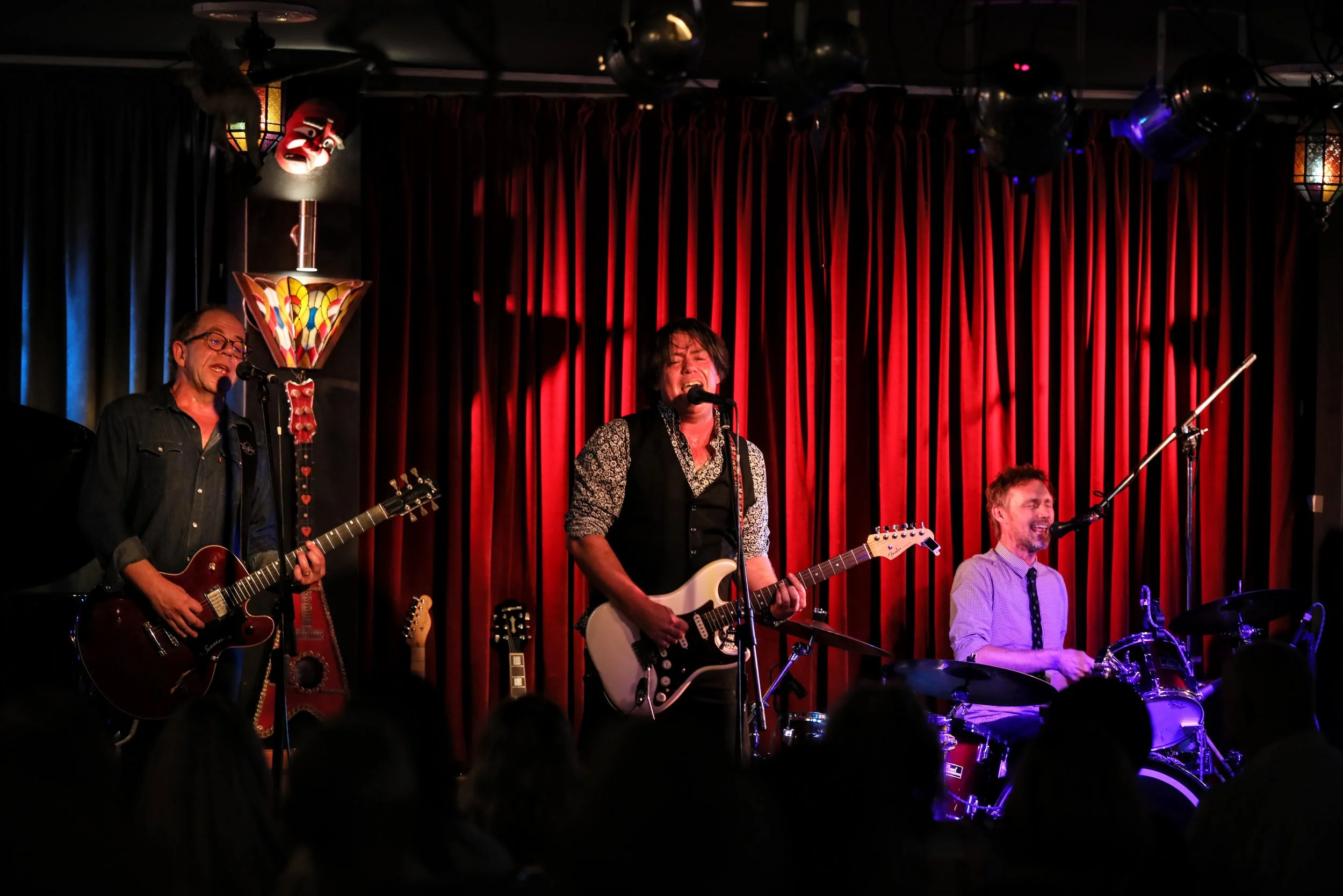 A live band performs on stage with a red curtain backdrop. The band includes three male musicians playing guitar, electric guitar, and drums, all singing into microphones. The stage is decorated with colorful lamps and spectators are visible in the f