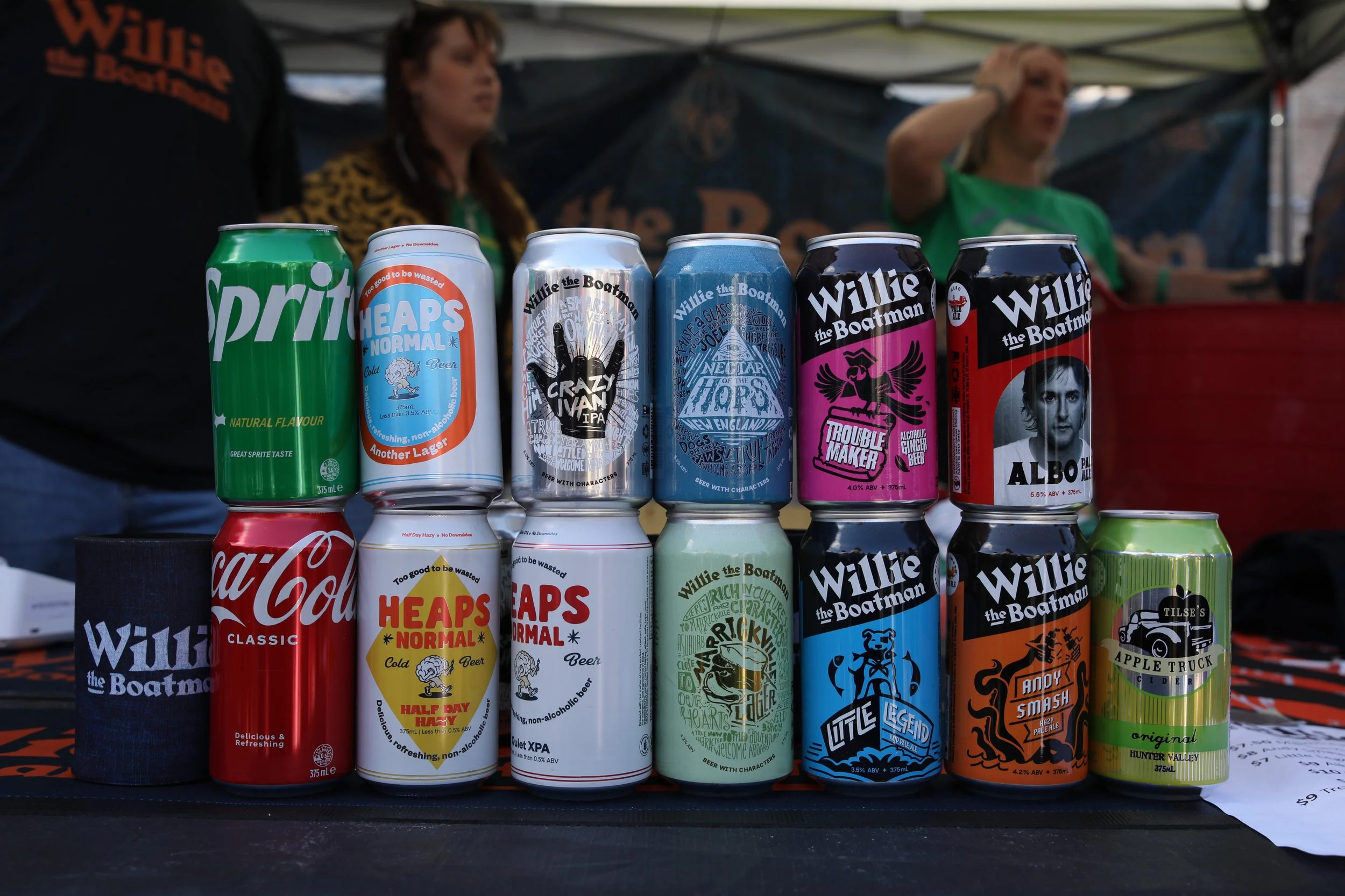 A variety of colorful canned beverages displayed on a table at an outdoor event, with people standing in the background under a tent.