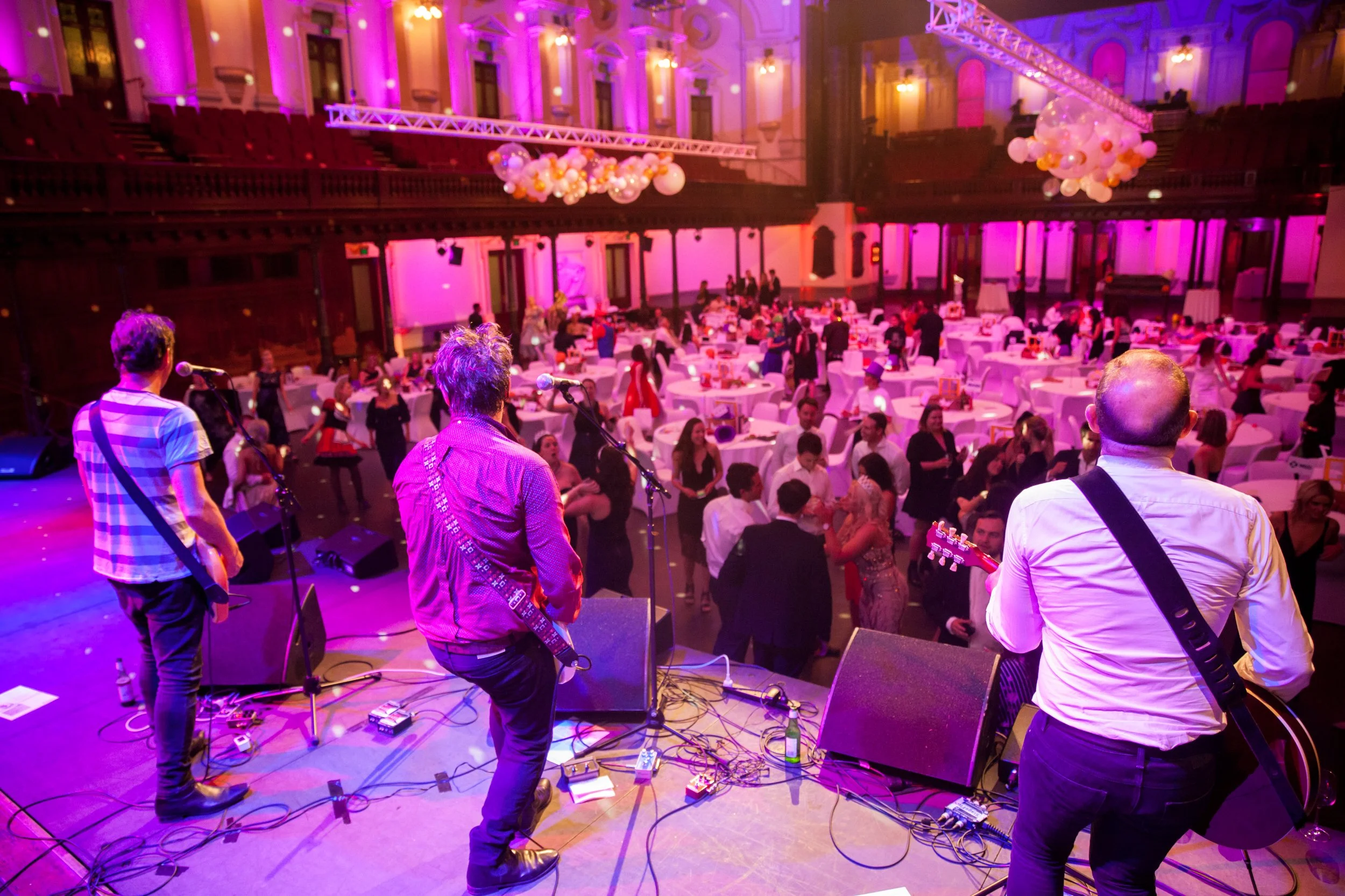 Band performing on stage at a wedding reception with purple and pink lighting, guests dancing and sitting at round tables with white tablecloths in a grand hall.