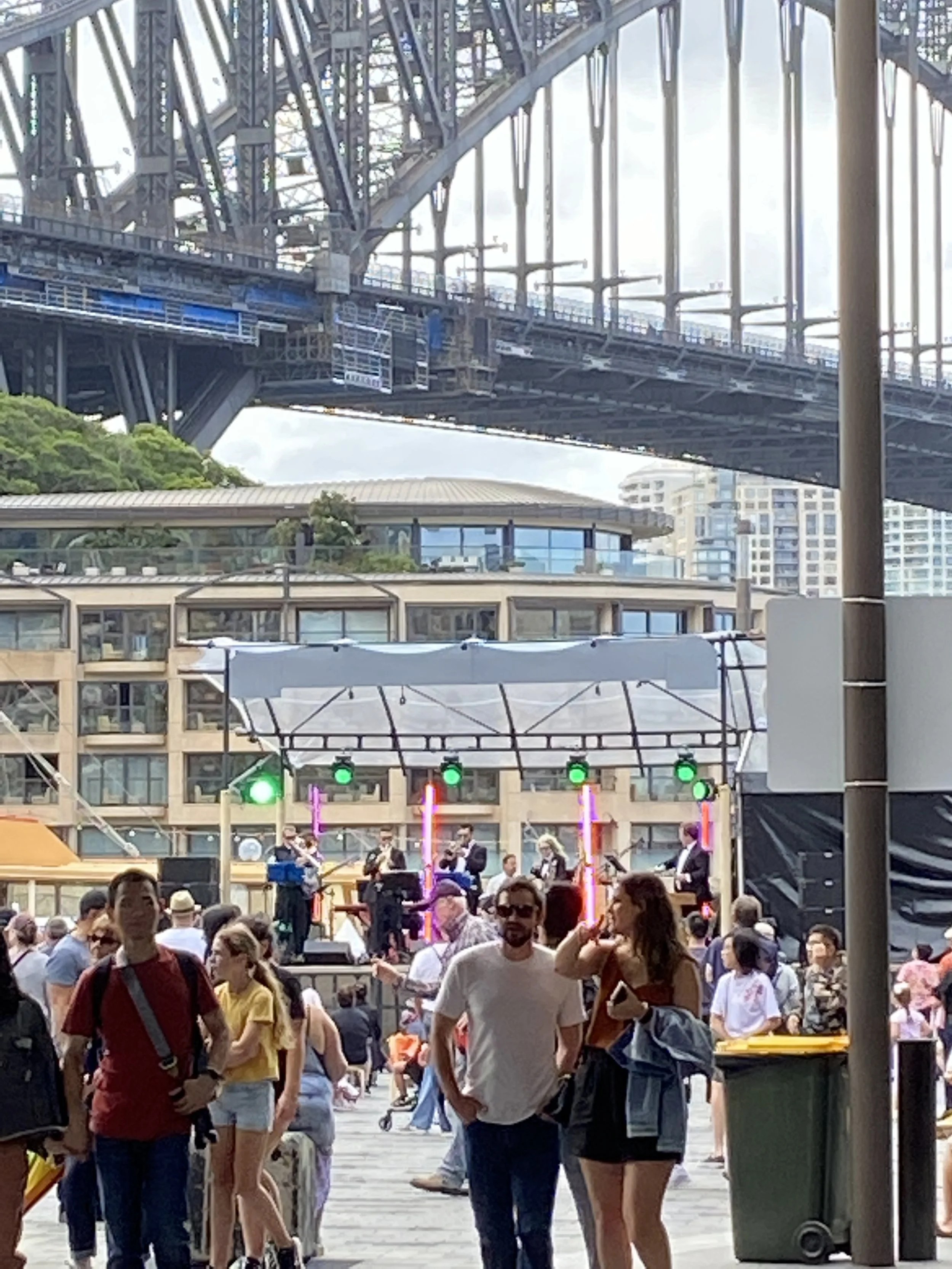 People attending an outdoor concert in a city, with a stage, musical band, and a bridge overhead.
