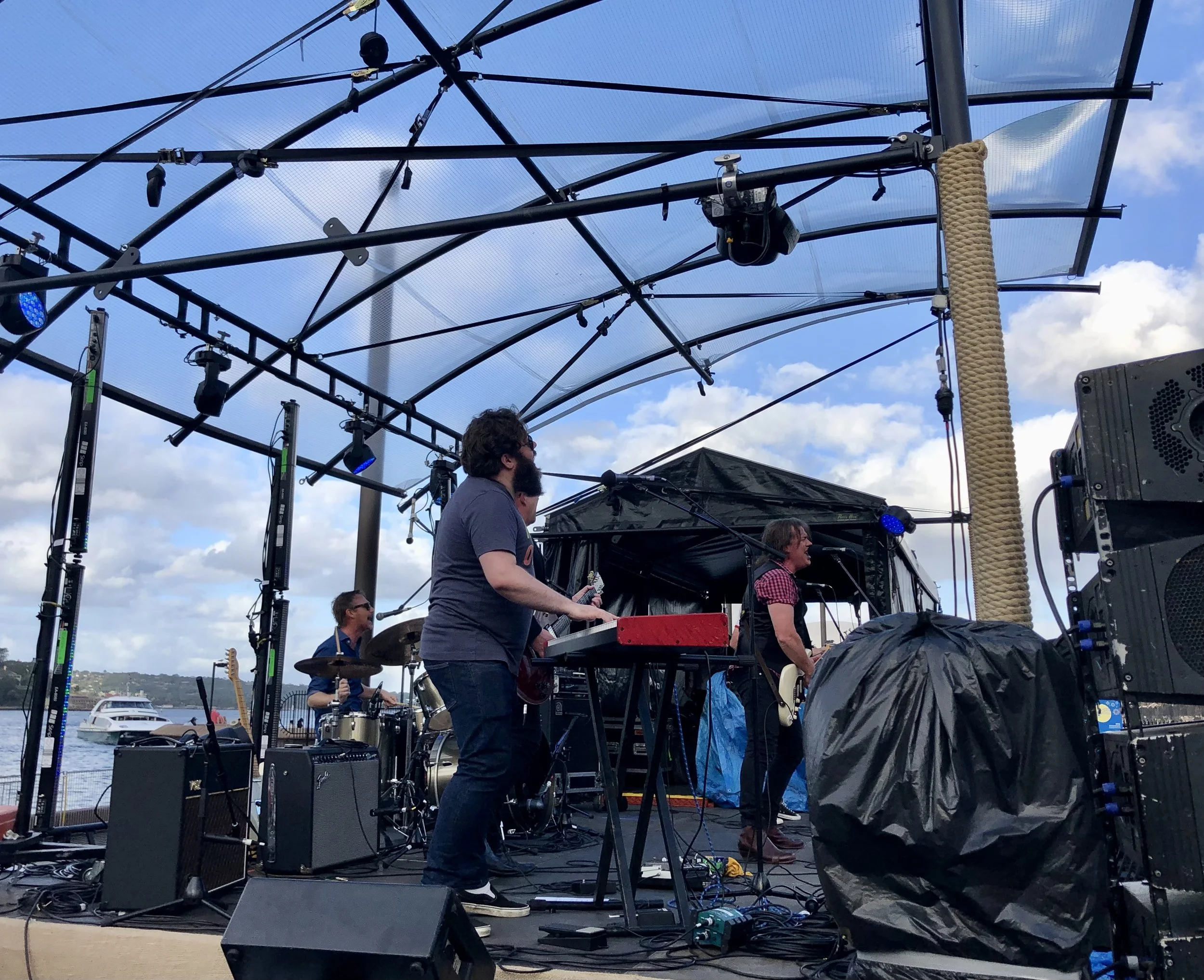 Band performing on outdoor stage with musical instruments and equipment, under a sky with clouds, near a body of water.