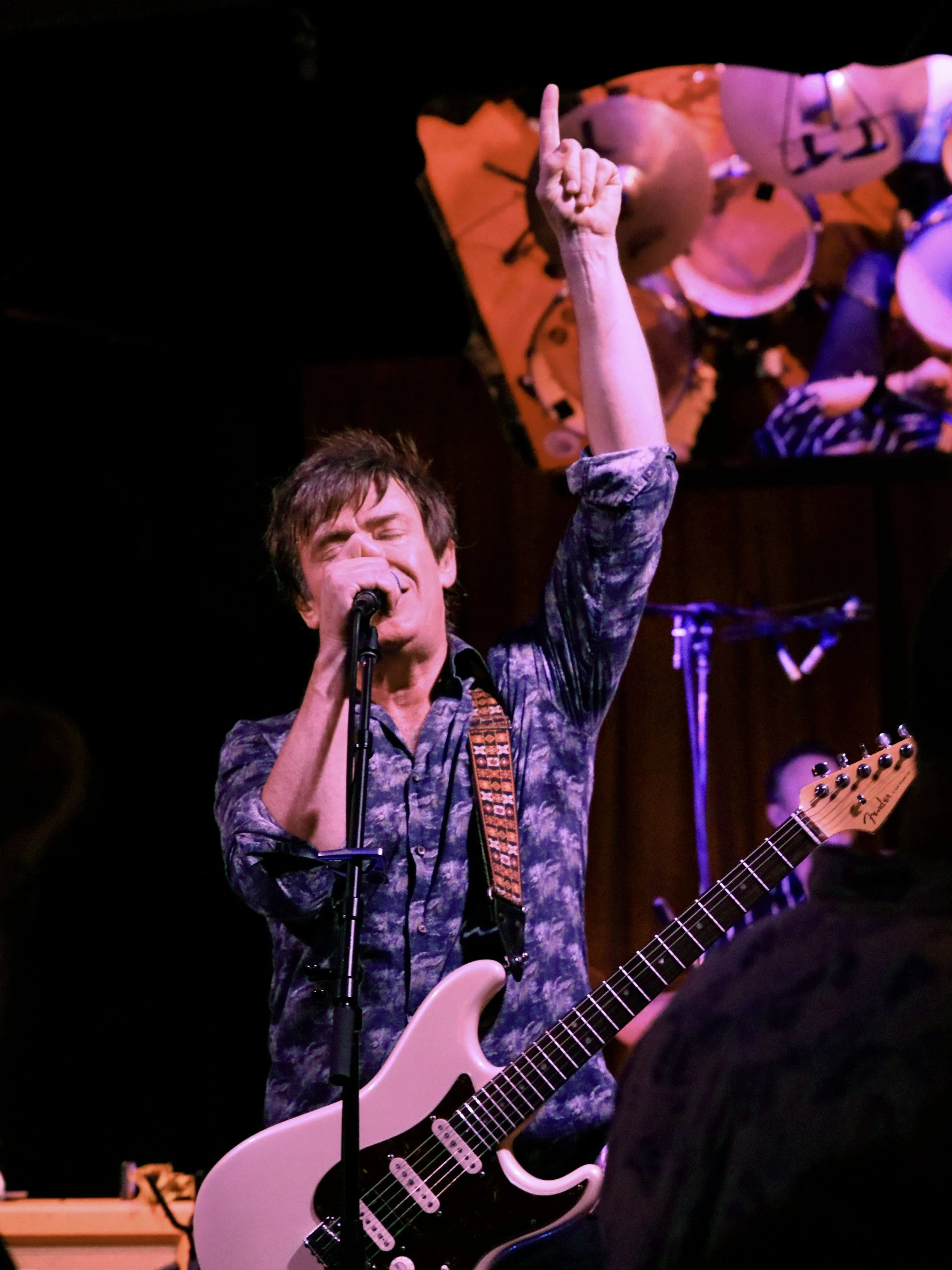 A man singing into a microphone while playing a white electric guitar, with his left hand raised and index finger pointing upwards, performing on stage.
