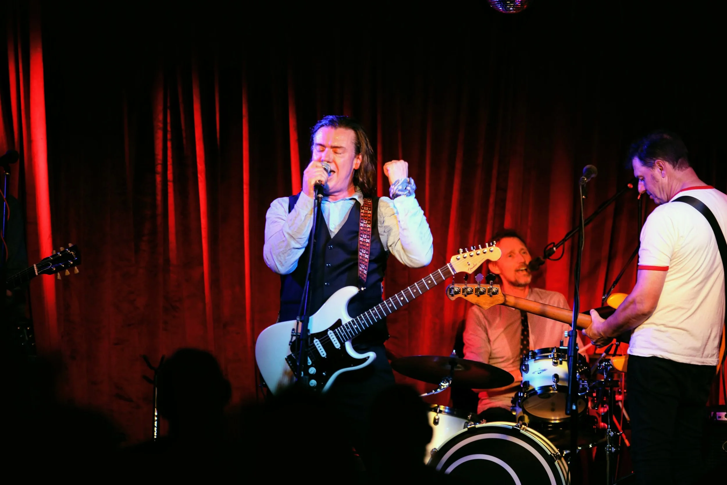 A band performing on stage with a lead singer playing guitar and singing into a microphone, a drummer in the background, and another guitarist to the right, all under red stage curtains.