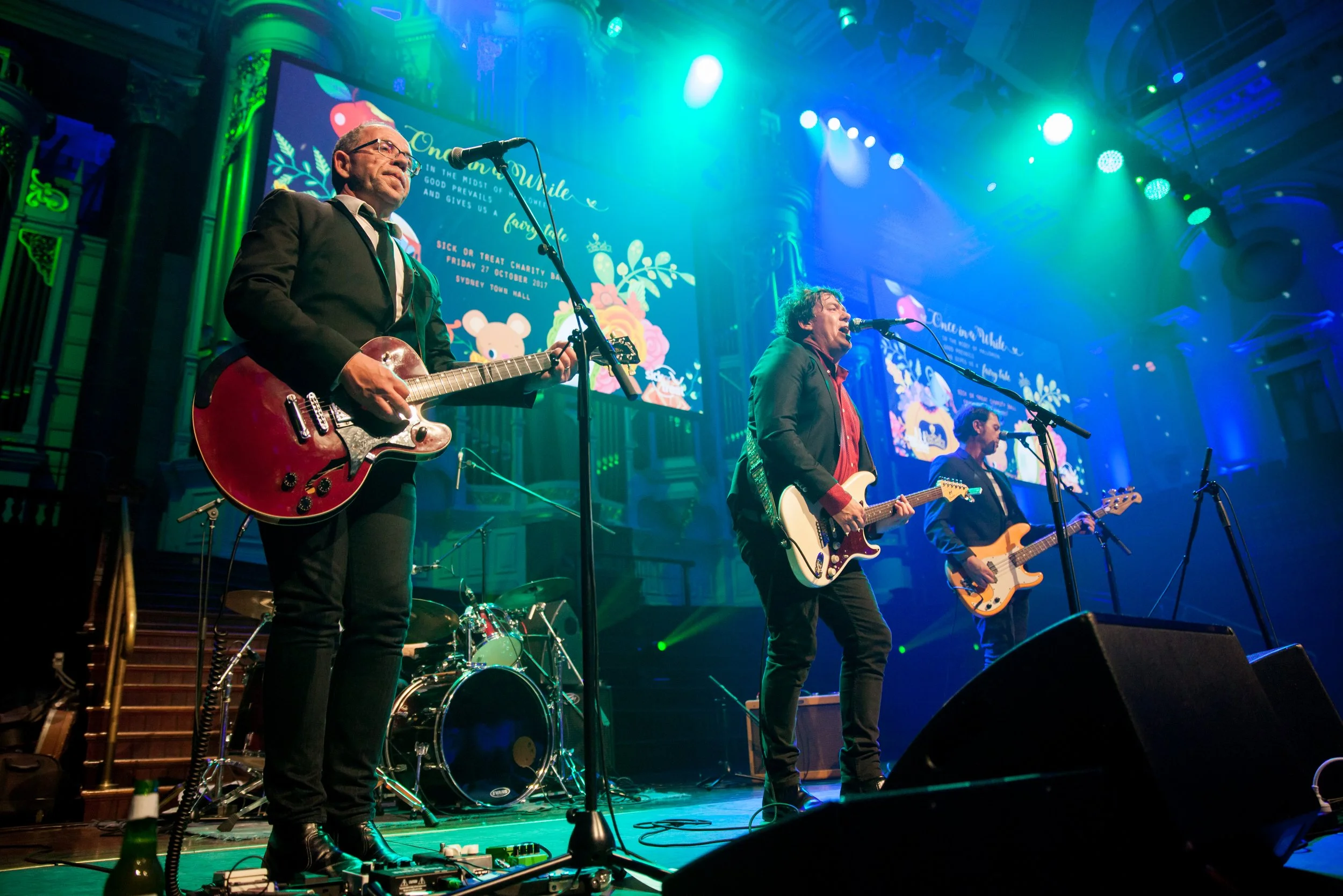 A band performing on stage with three musicians playing guitars and drums in the background, illuminated by colorful stage lights.