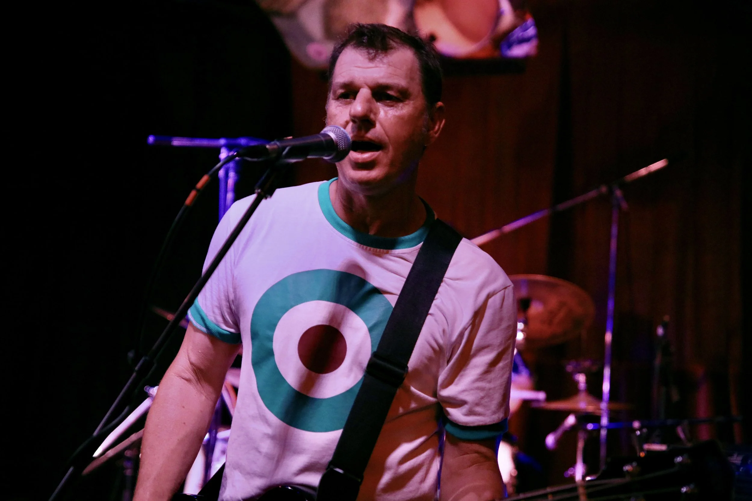Male musician singing into a microphone while playing guitar on stage, with drum set in the background, in a dimly lit venue.