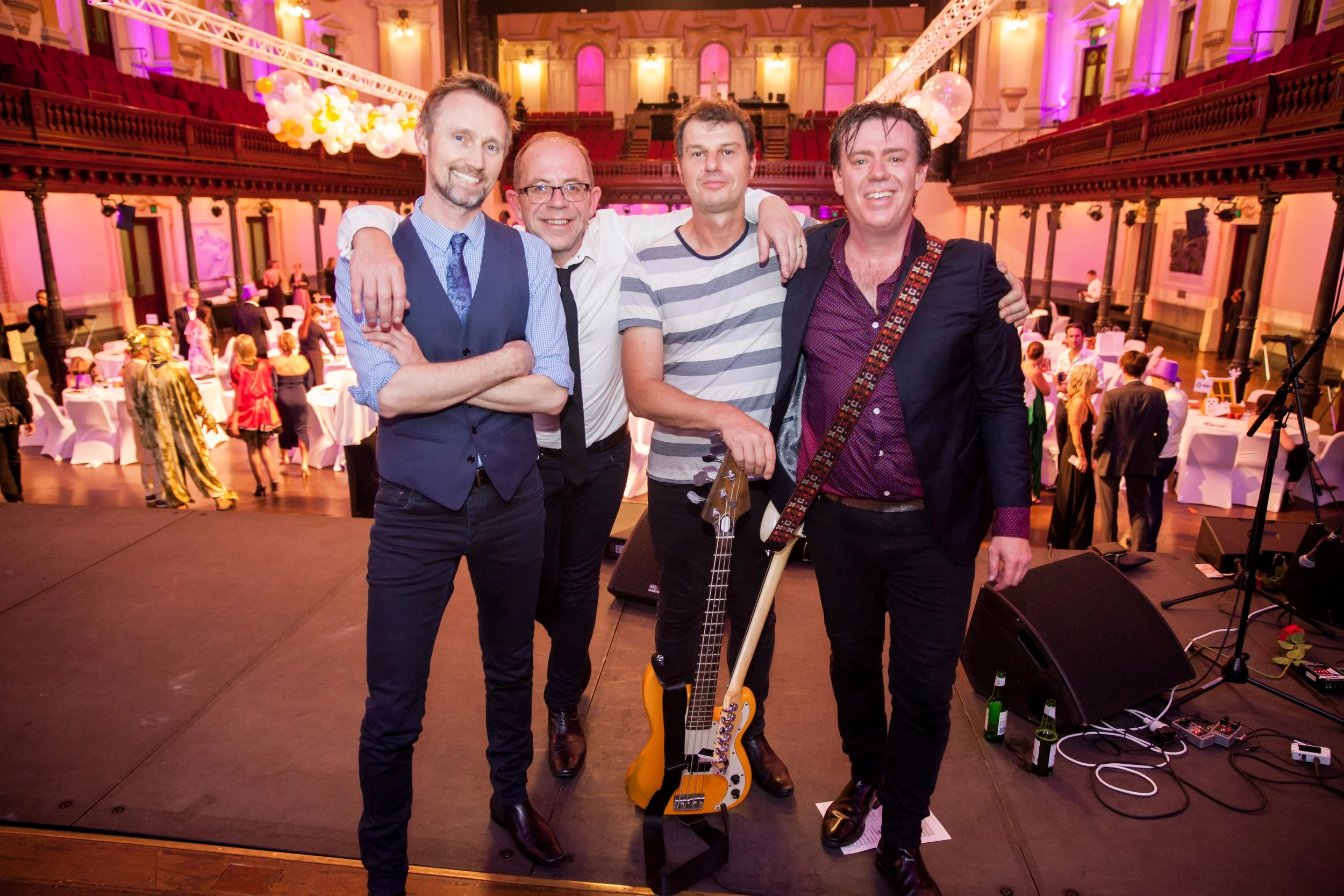 A band of four men standing close together on a stage at a social event in a large hall with round tables, balloons, and pink lighting. One man is holding a guitar, and all are smiling at the camera.