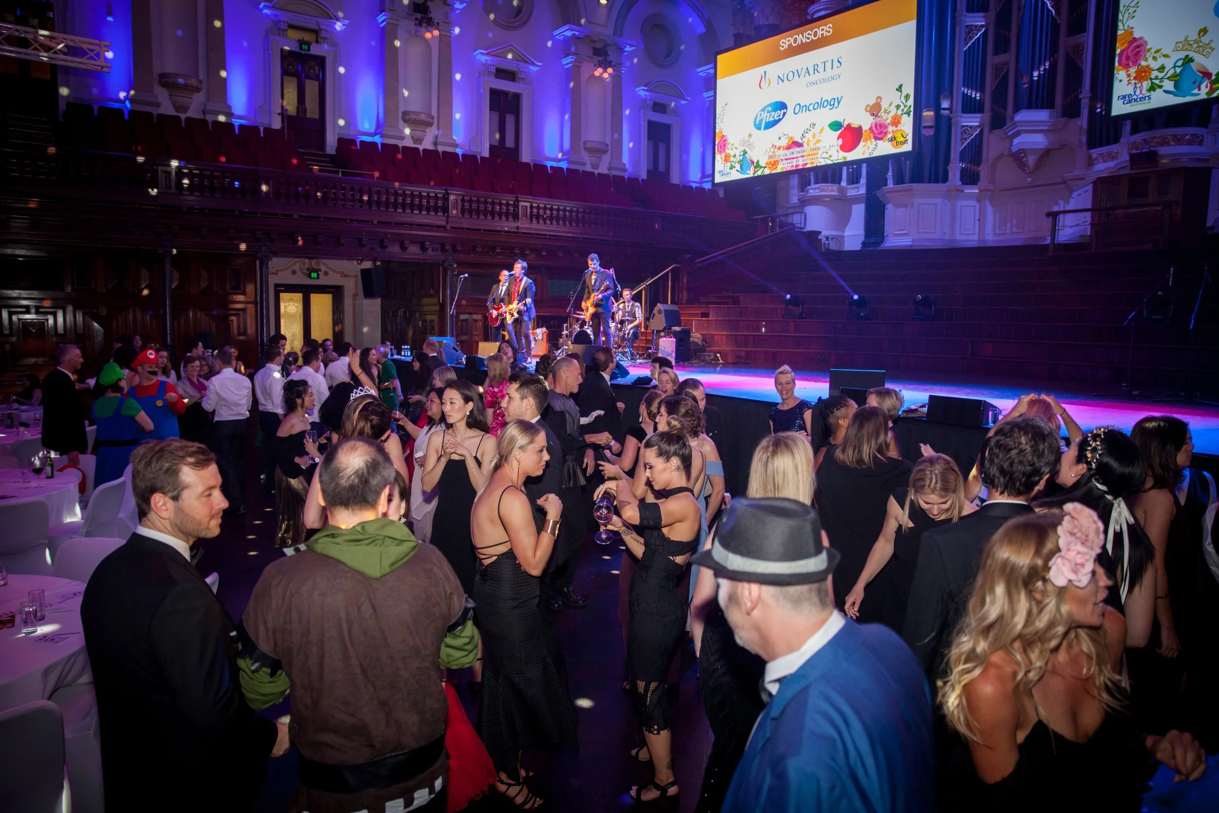 Guests at a lively indoor event dance near stage where a band performs, with a large screen displaying sponsors in the background.