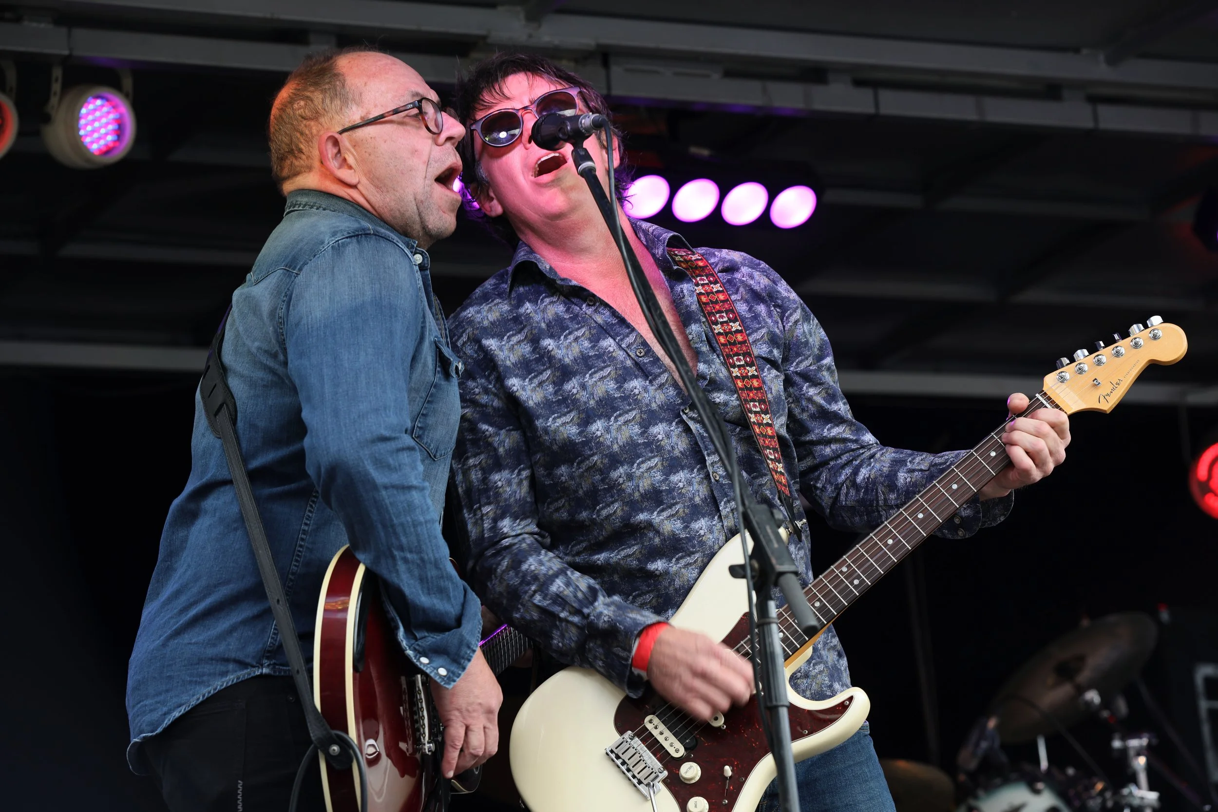 Two male musicians performing on stage with electric guitars. The man on the left is wearing glasses and a denim shirt, and the man on the right is wearing sunglasses and a patterned shirt. They are singing into a microphone together.