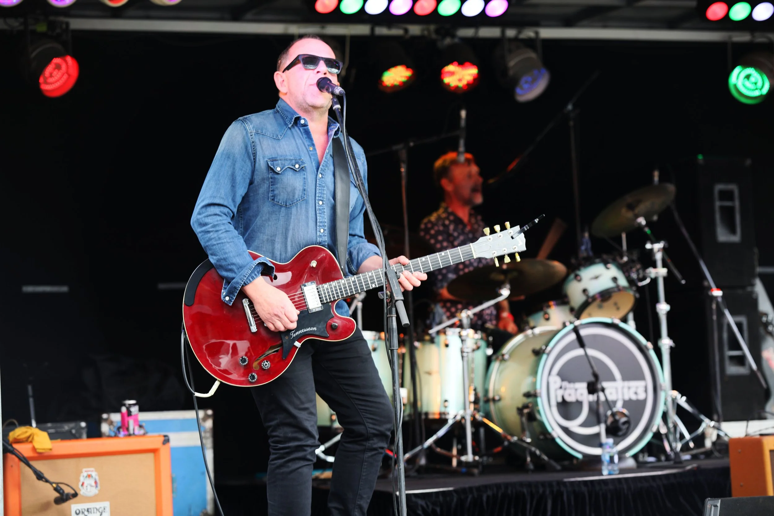 Musical performance featuring a male singer and guitarist in sunglasses and denim shirt, with a band playing drums on stage under colorful lights.