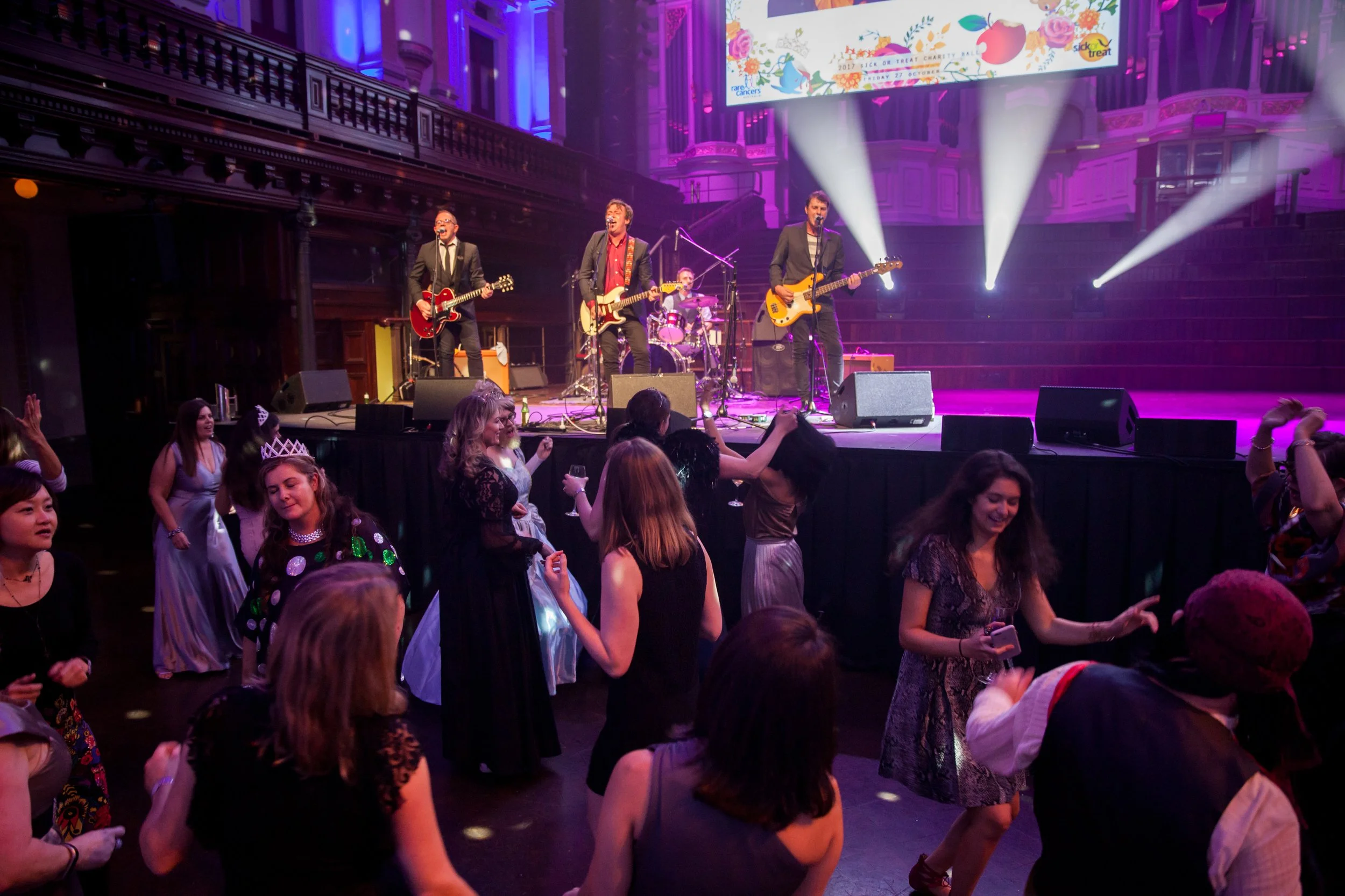 People dancing and enjoying live music performance at an indoor concert with bright stage lights and a band playing on stage.