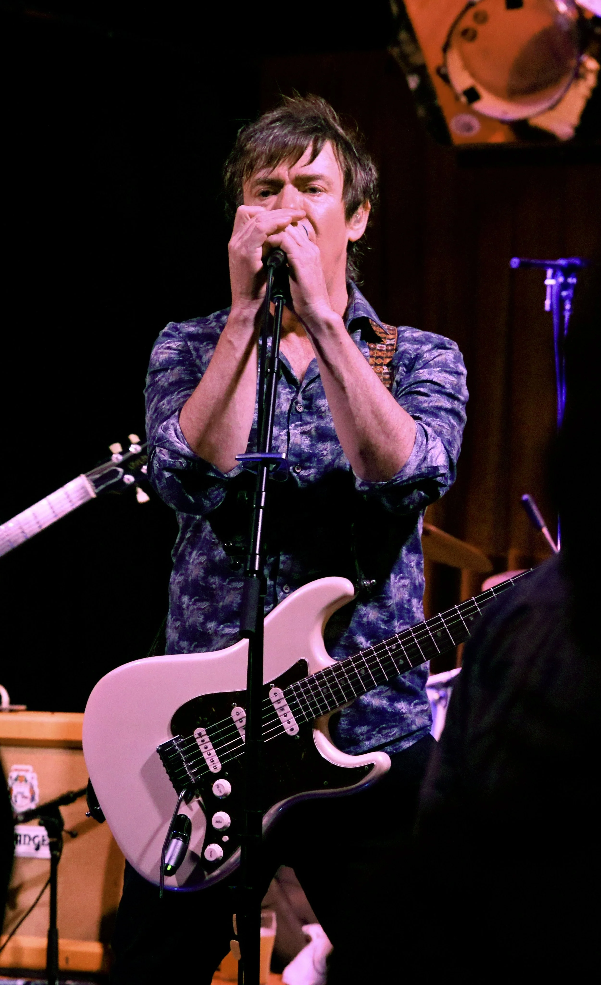 Musician with brown hair playing a pink electric guitar and singing into a microphone on stage.