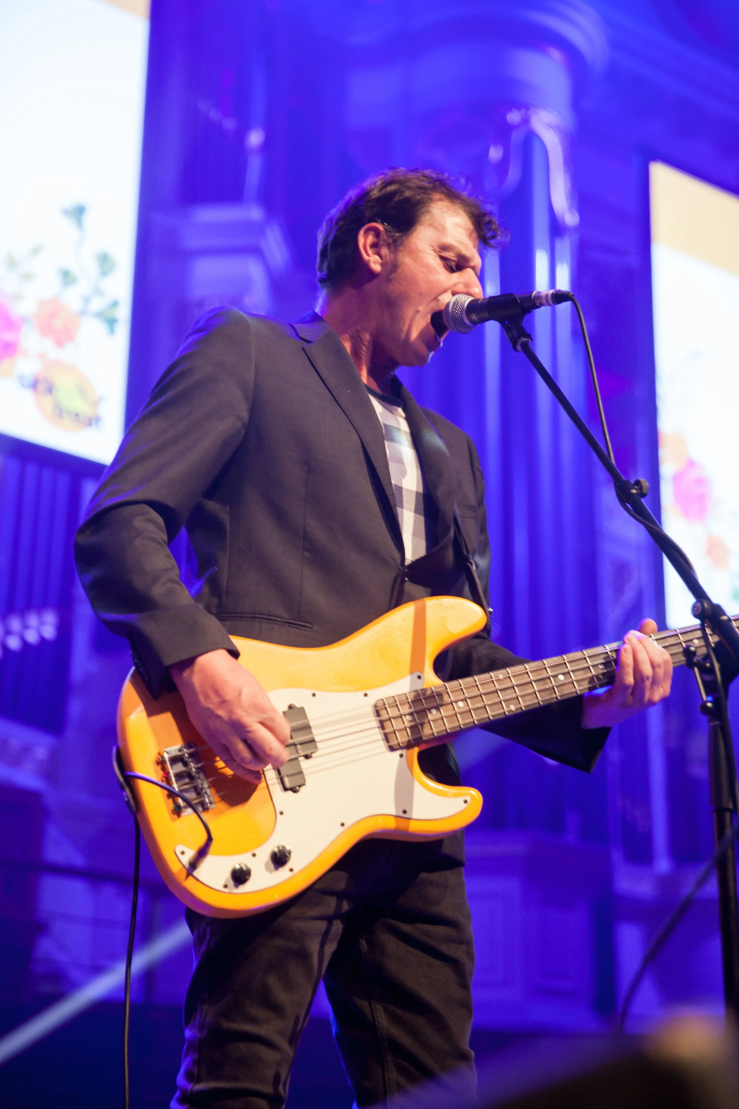 A man in a jacket performs on stage playing an electric guitar and singing into a microphone with purple lighting and a large screen in the background.