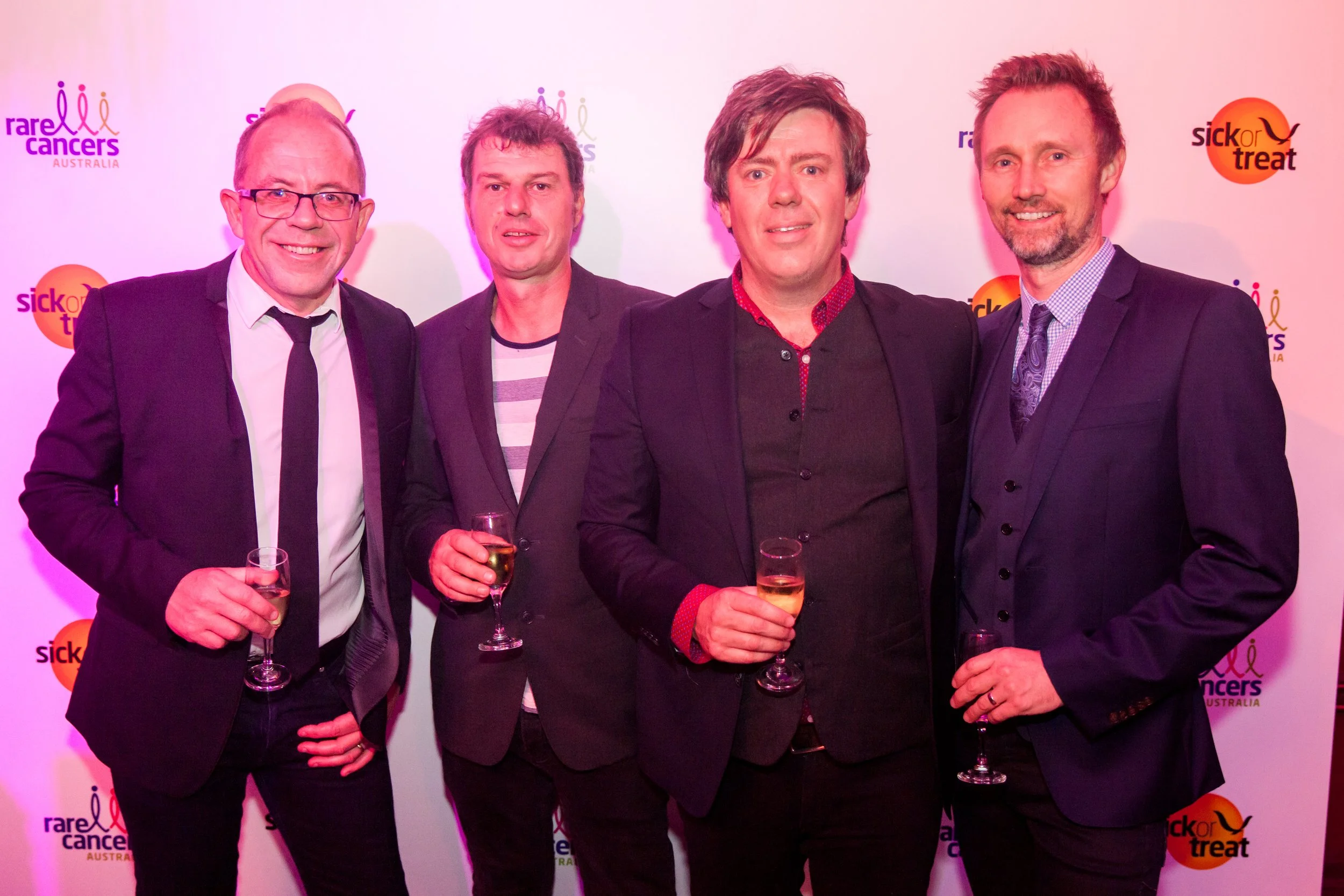 Four men in suits and casual attire standing at an event, holding glasses of champagne, with a backdrop featuring logos for Rare Cancers Australia and Sick for Treet.
