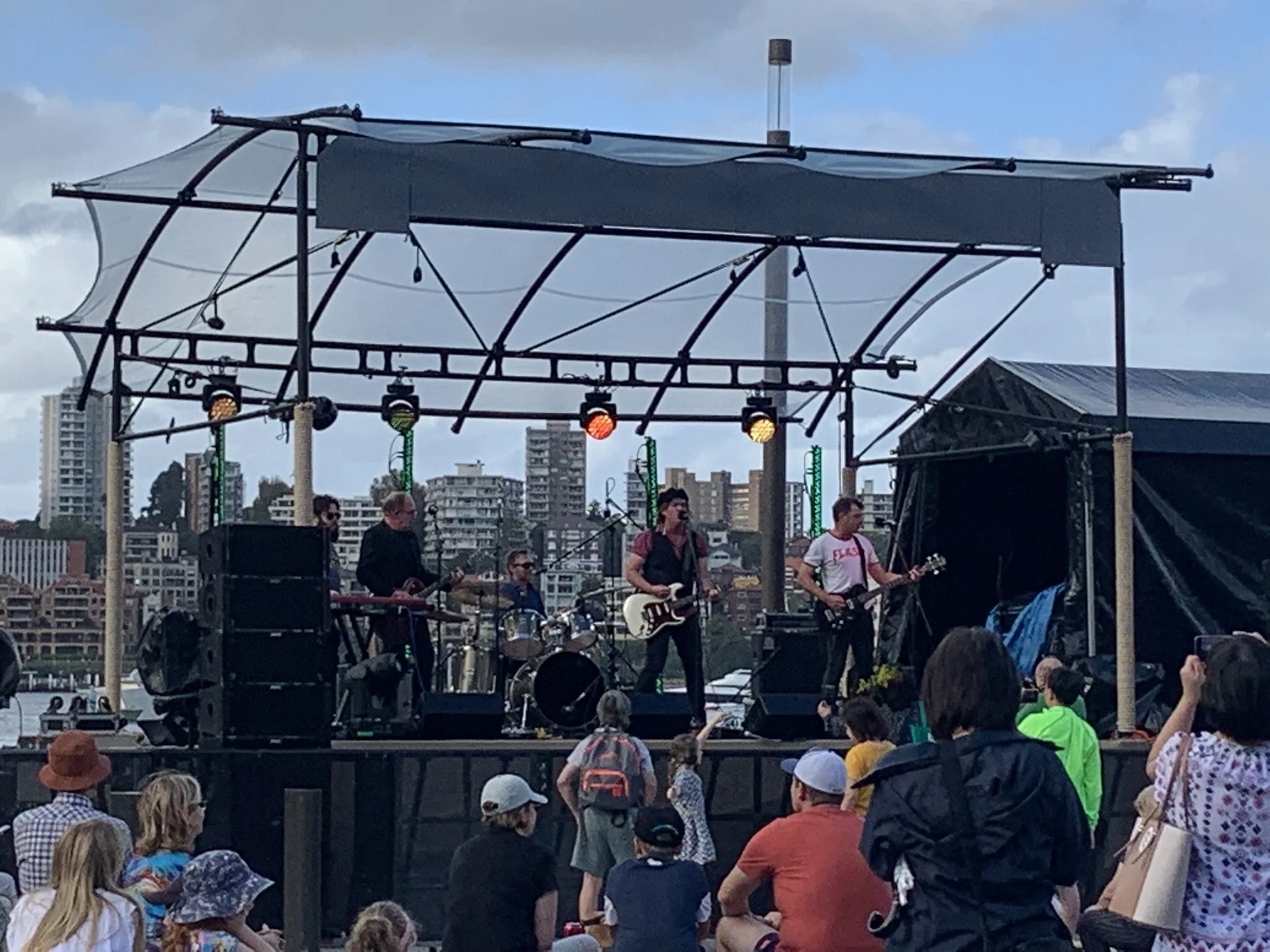 A live outdoor concert with a band performing on a stage, audience members watching, some wearing hats and jackets, under a cloudy sky in a cityscape setting.