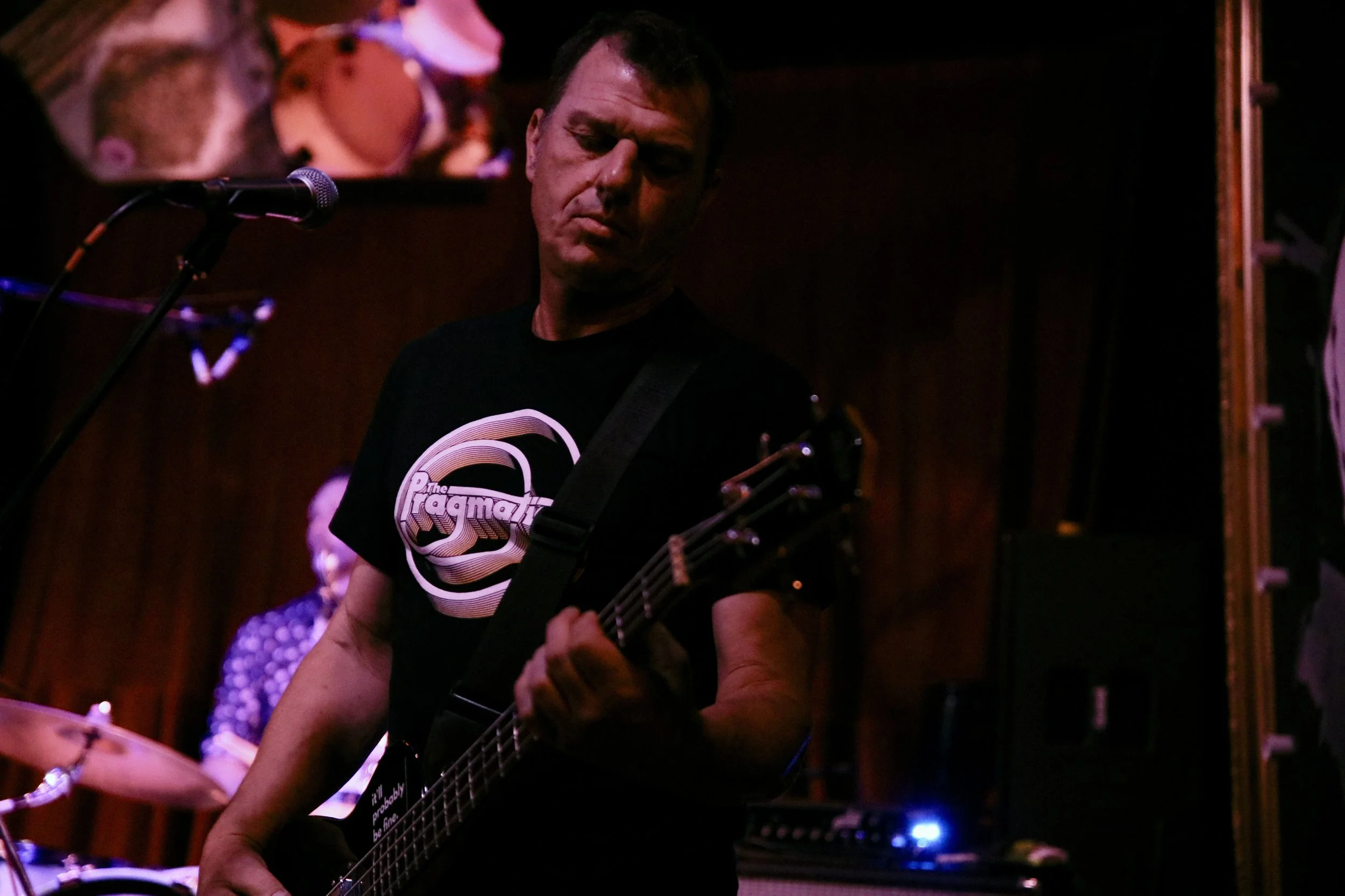 A man playing an electric guitar during a live performance, with a woman playing drums in the background, in a dimly lit venue.