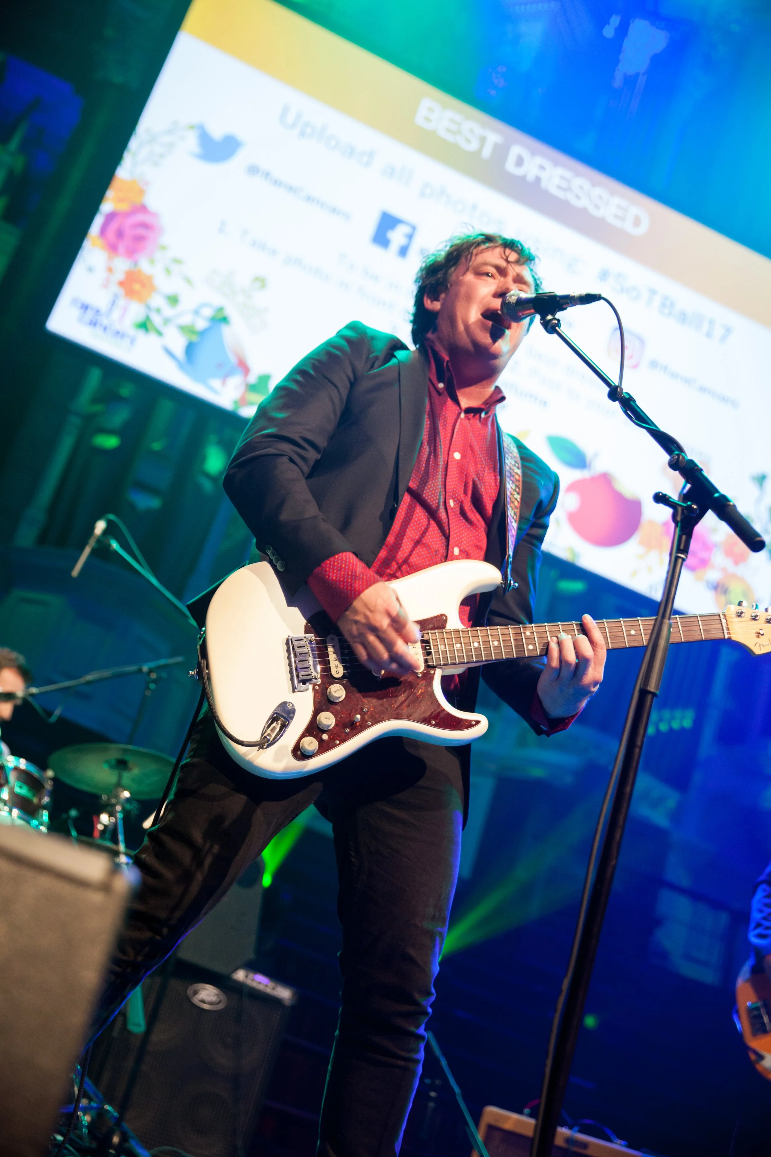 Man playing electric guitar and singing into a microphone on stage at an event, with a colorful screen behind him displaying text about social media.