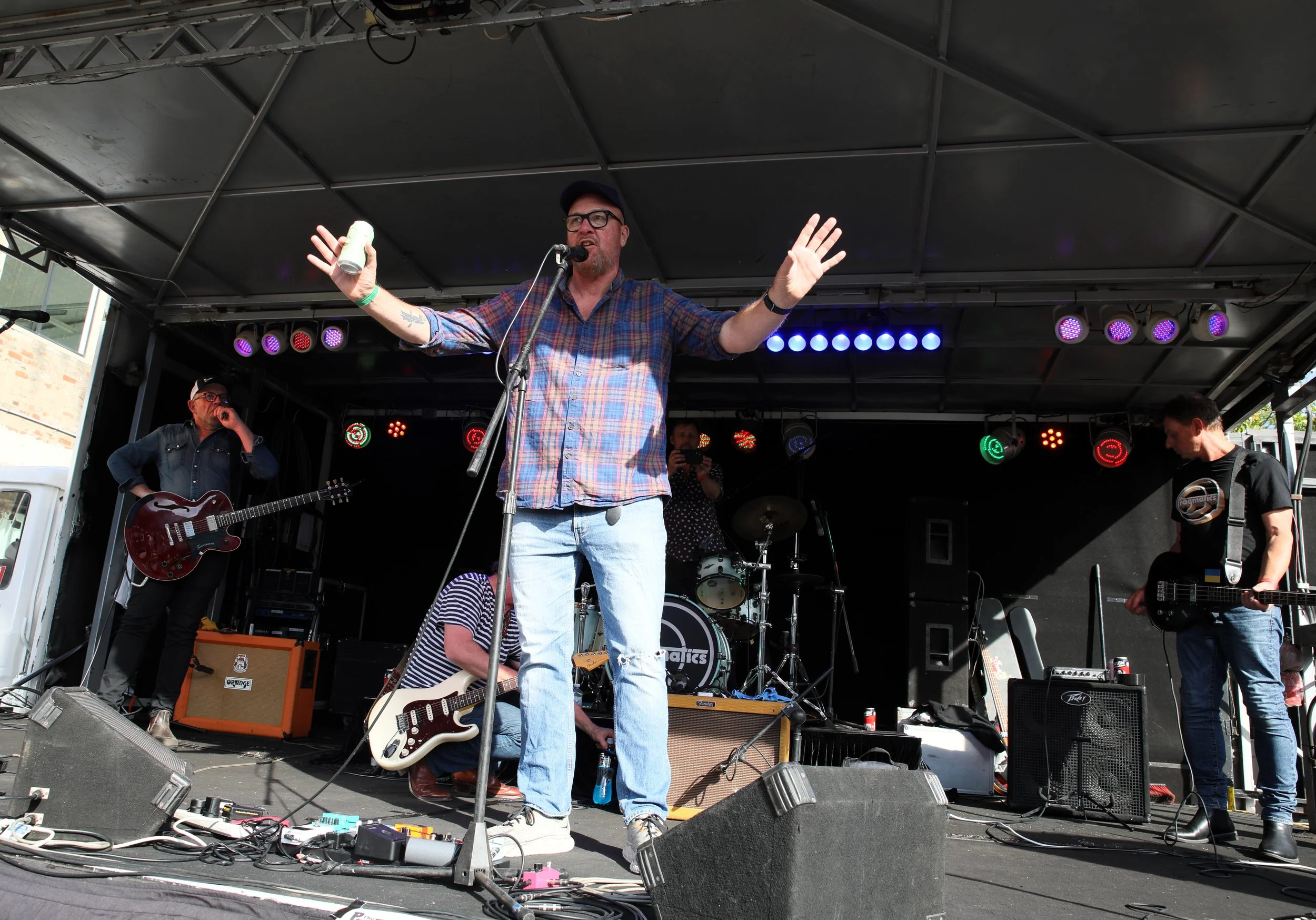 Band performing live on stage with lead singer in plaid shirt and light jeans, three musicians playing guitar and drums, colorful stage lights.