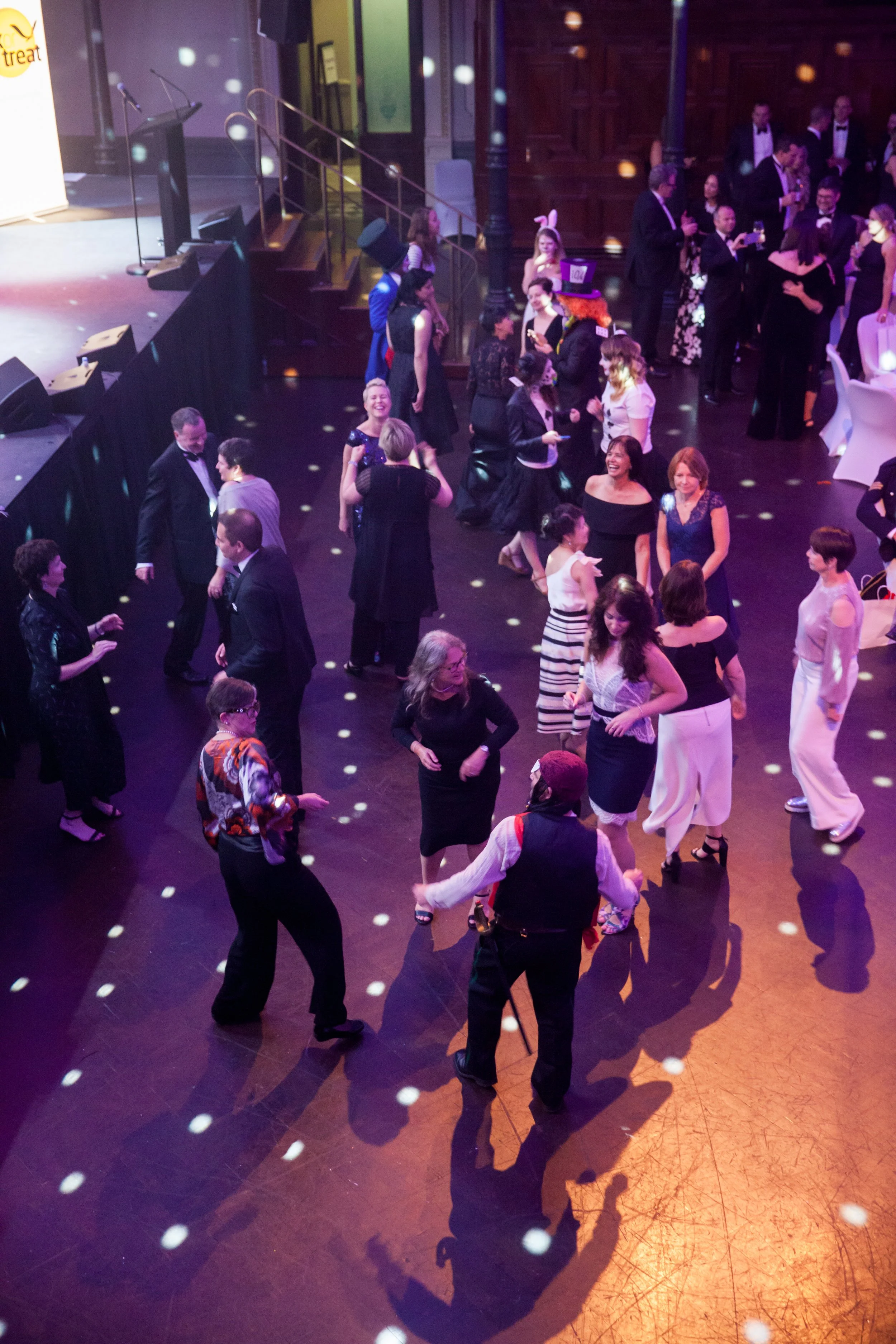 People dancing and socializing at a formal event with a stage and a podium in the background, some wearing tuxedos and gowns, others in casual elegant attire, with colorful lighting and disco effects.