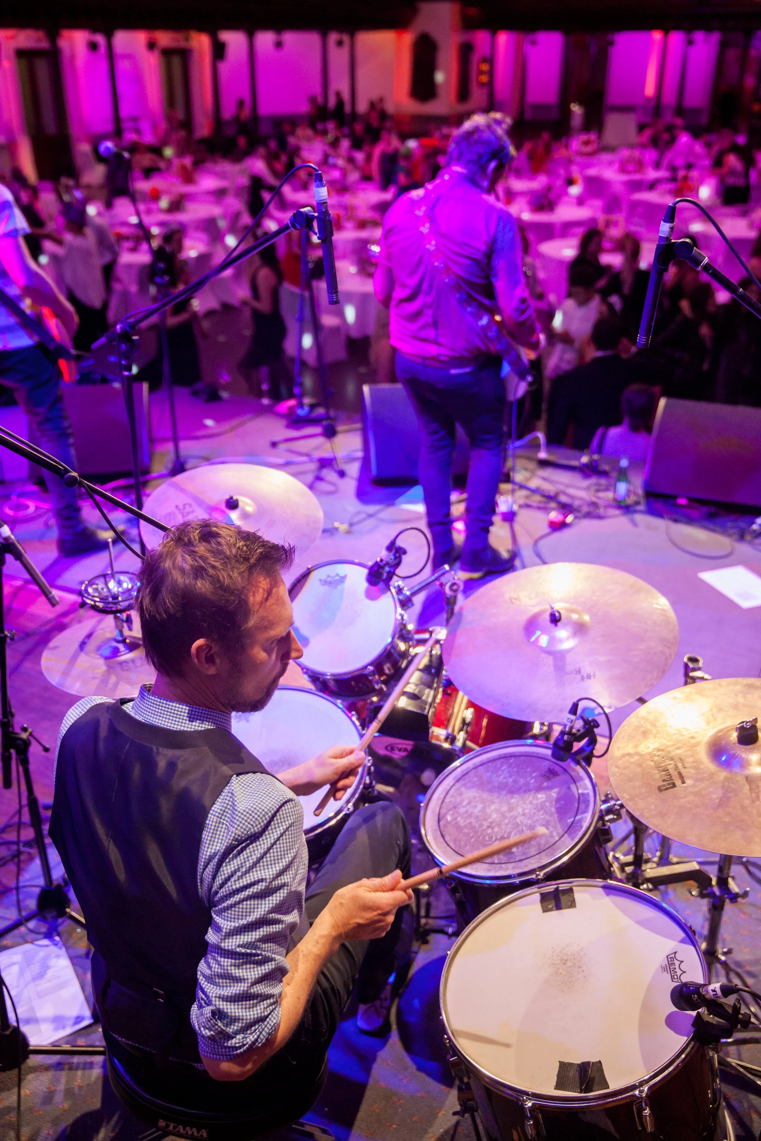 Musicians performing live on stage at a concert or event, with a drummer in the foreground and a guitarist in the background, and a lively audience seated at tables in a dimly lit venue with purple and pink lighting.