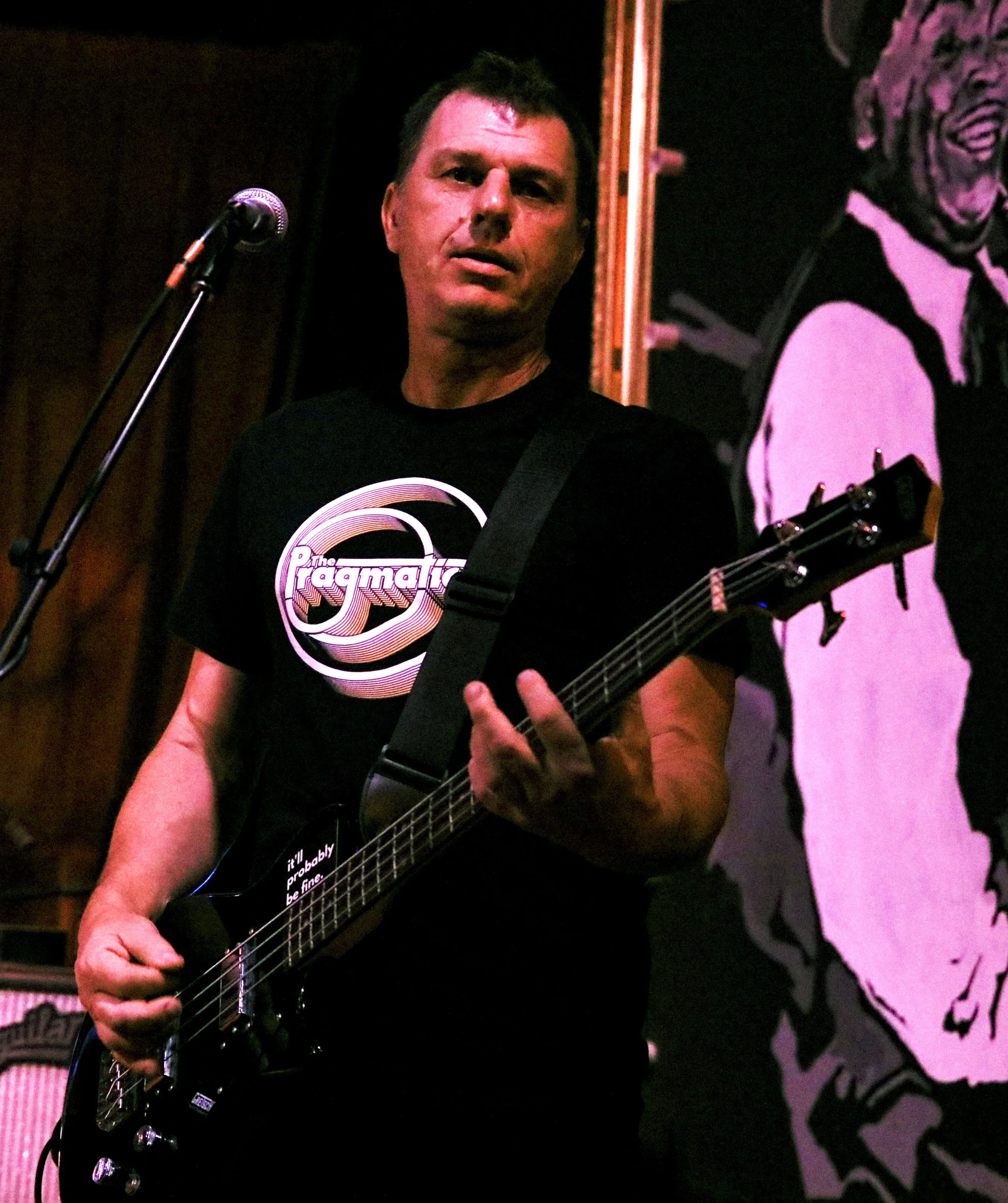 A man playing an electric guitar on stage, wearing a black t-shirt with pink and white logo and standing in front of a black background with a large illustrated poster of a laughing man.